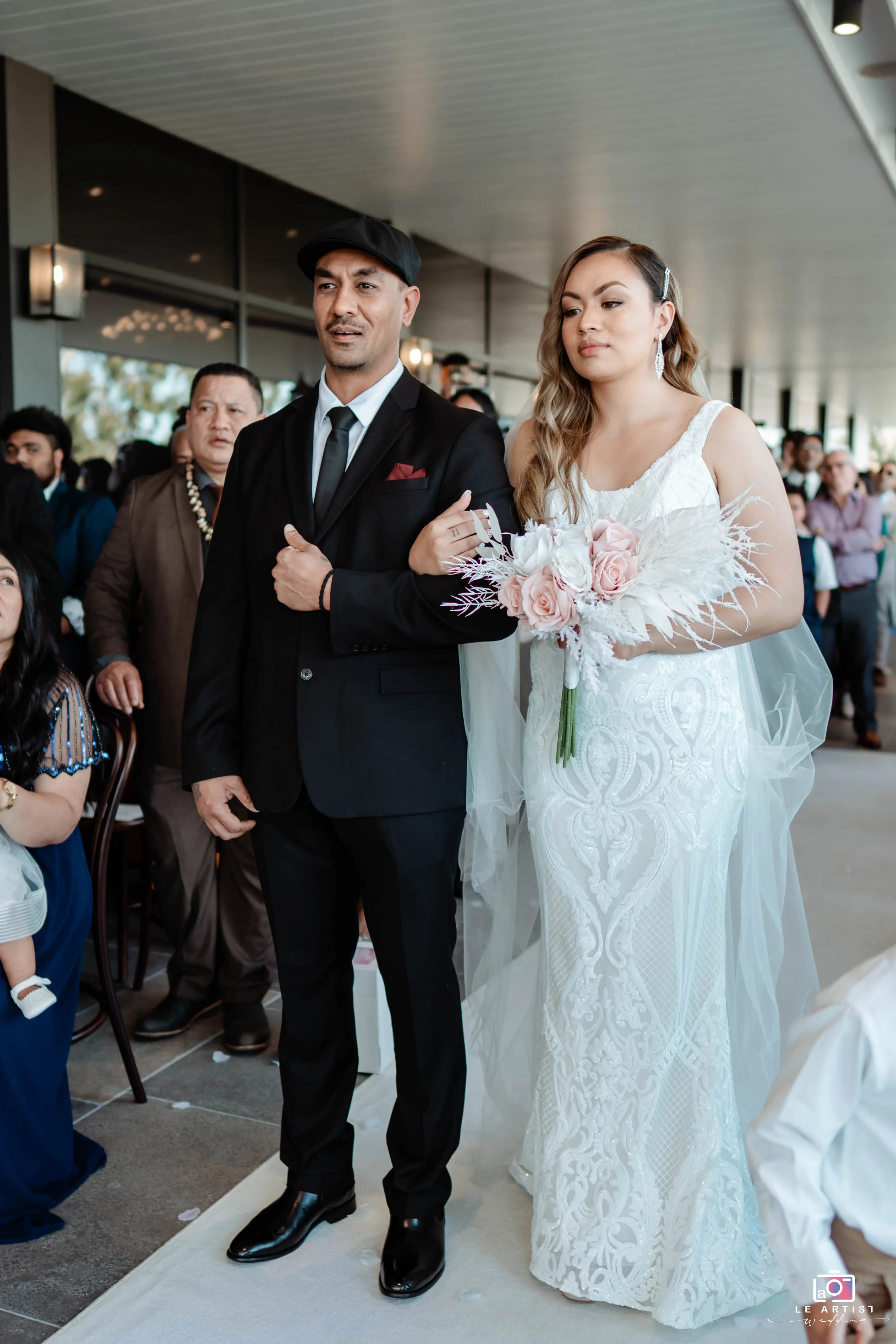 The bride walks down the aisle, arm in arm with a companion, holding a bouquet of roses and adorned in a beautiful lace gown, as guests look on. Le Artist Wedding