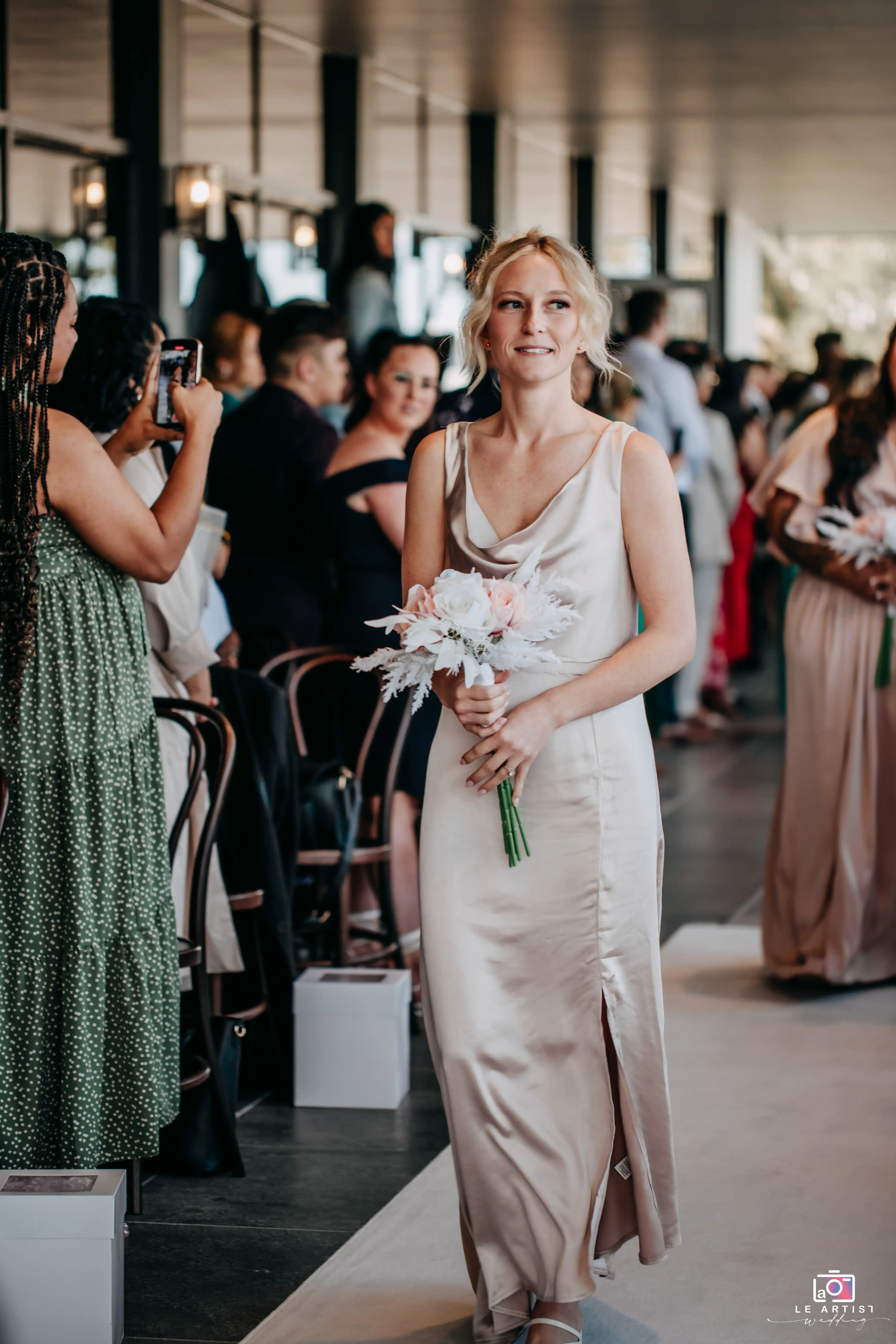 A bridesmaid gracefully walks down the aisle holding a bouquet, as guests capture the moment. Le Artist Wedding