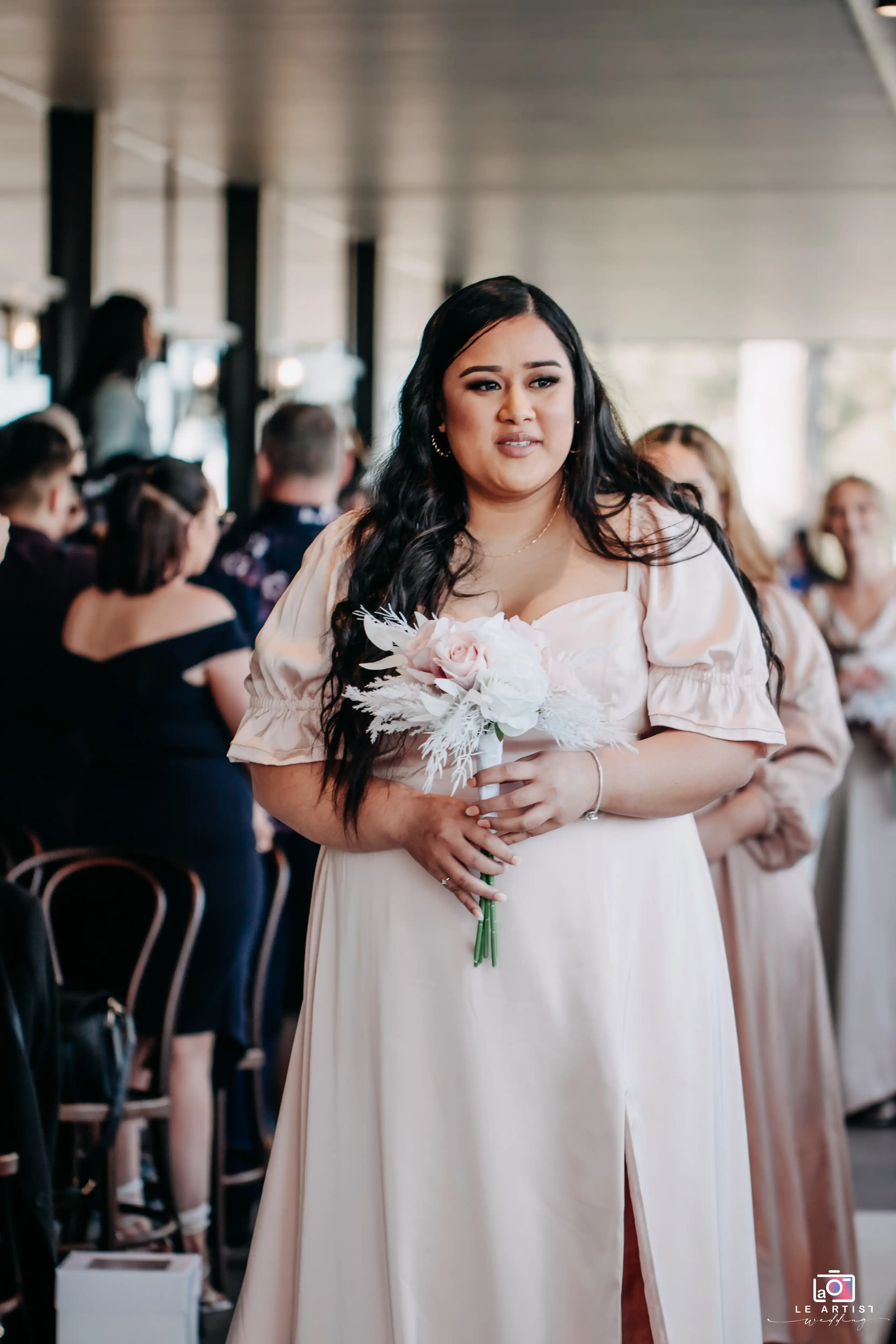 A bridesmaid walks down the aisle holding a delicate bouquet of pink and white flowers during the wedding processional. Le Artist Wedding