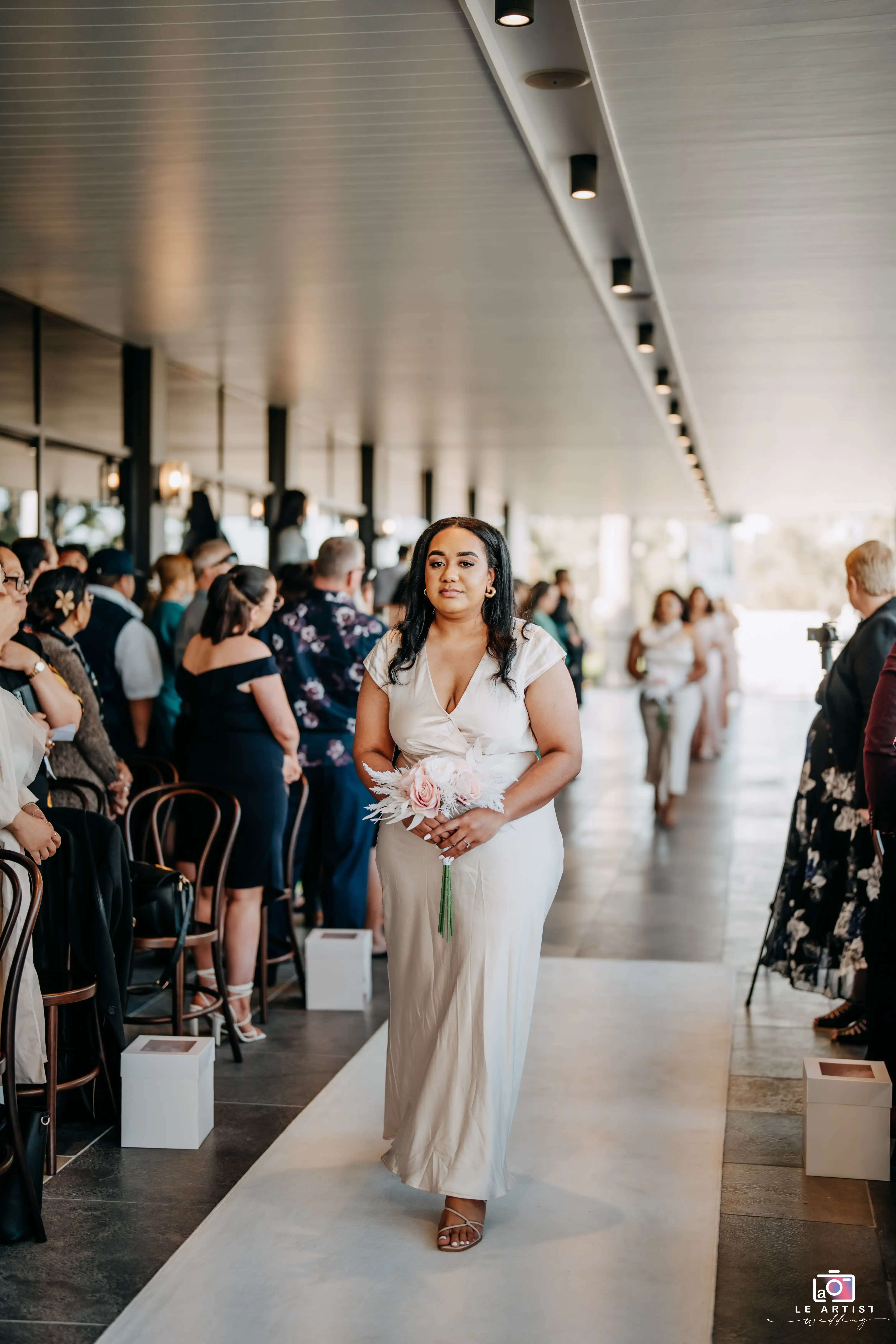 A bridesmaid gracefully walks down the aisle, holding a bouquet, as guests look on at the beautifully decorated venue. Le Artist Wedding