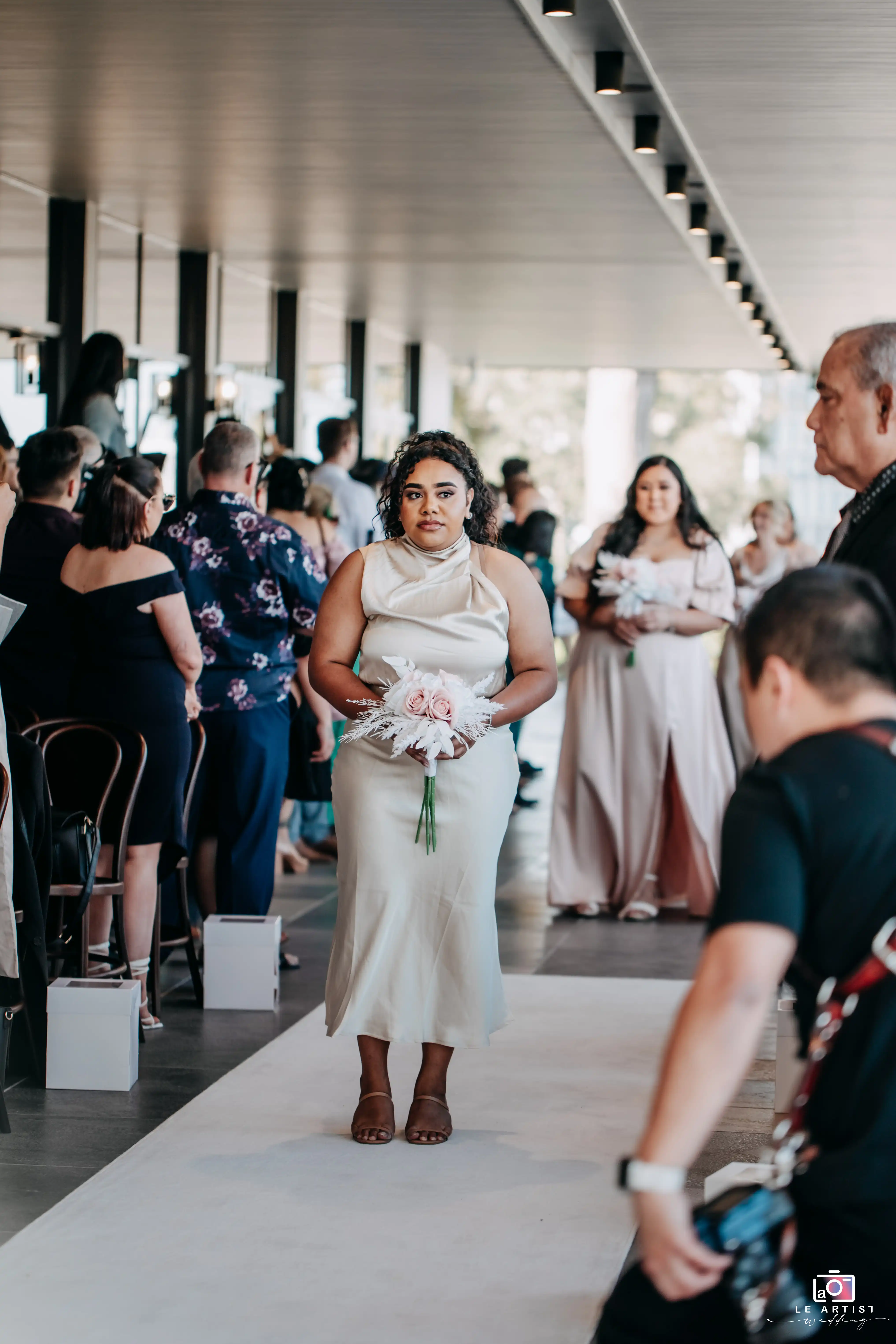 A bridesmaid walks down the aisle holding a pastel bouquet, marking the start of the ceremony processional. Le Artist Wedding