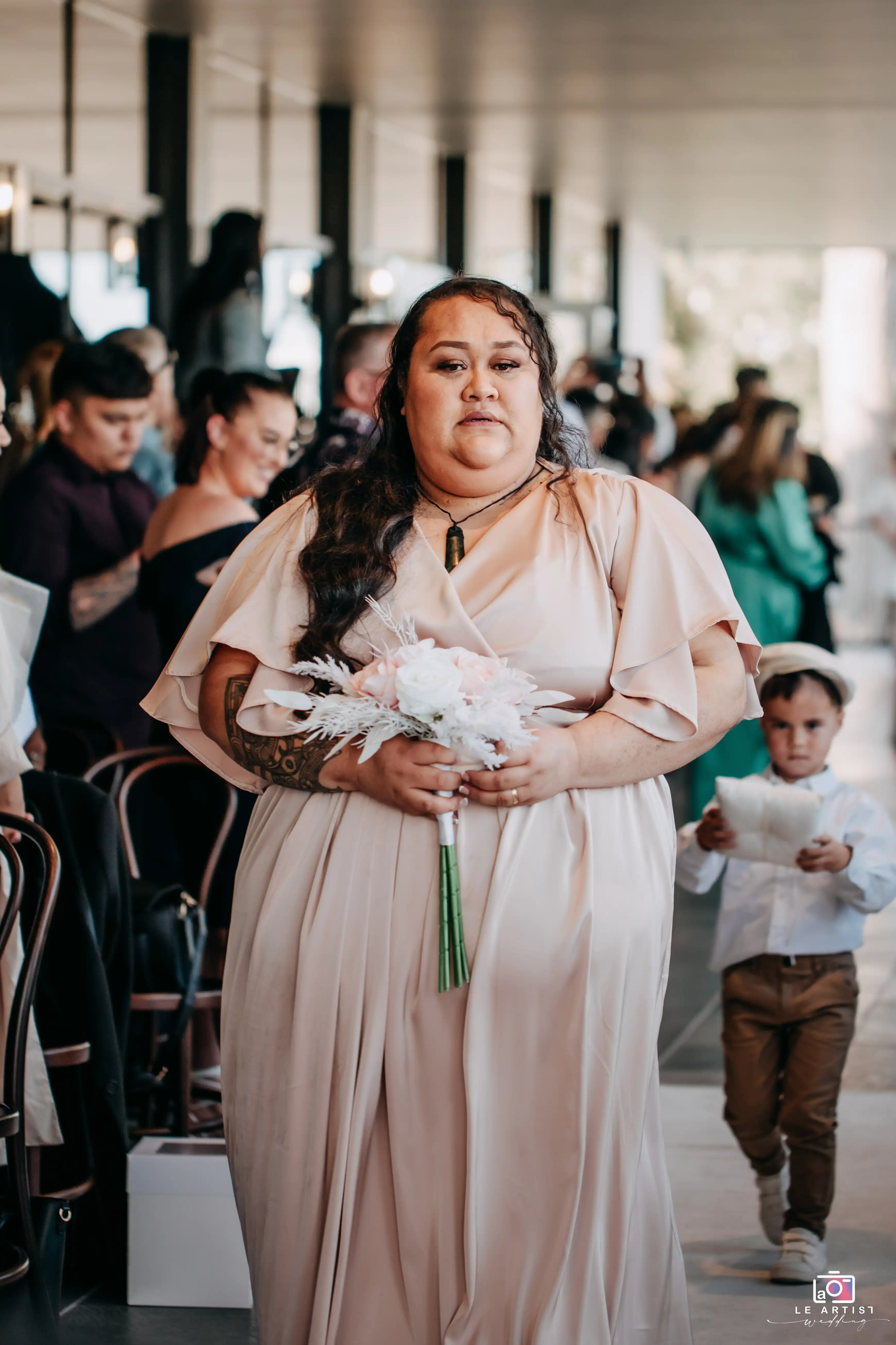 A bridesmaid walks down the aisle holding a bouquet, followed closely by a young ring bearer. Le Artist Wedding
