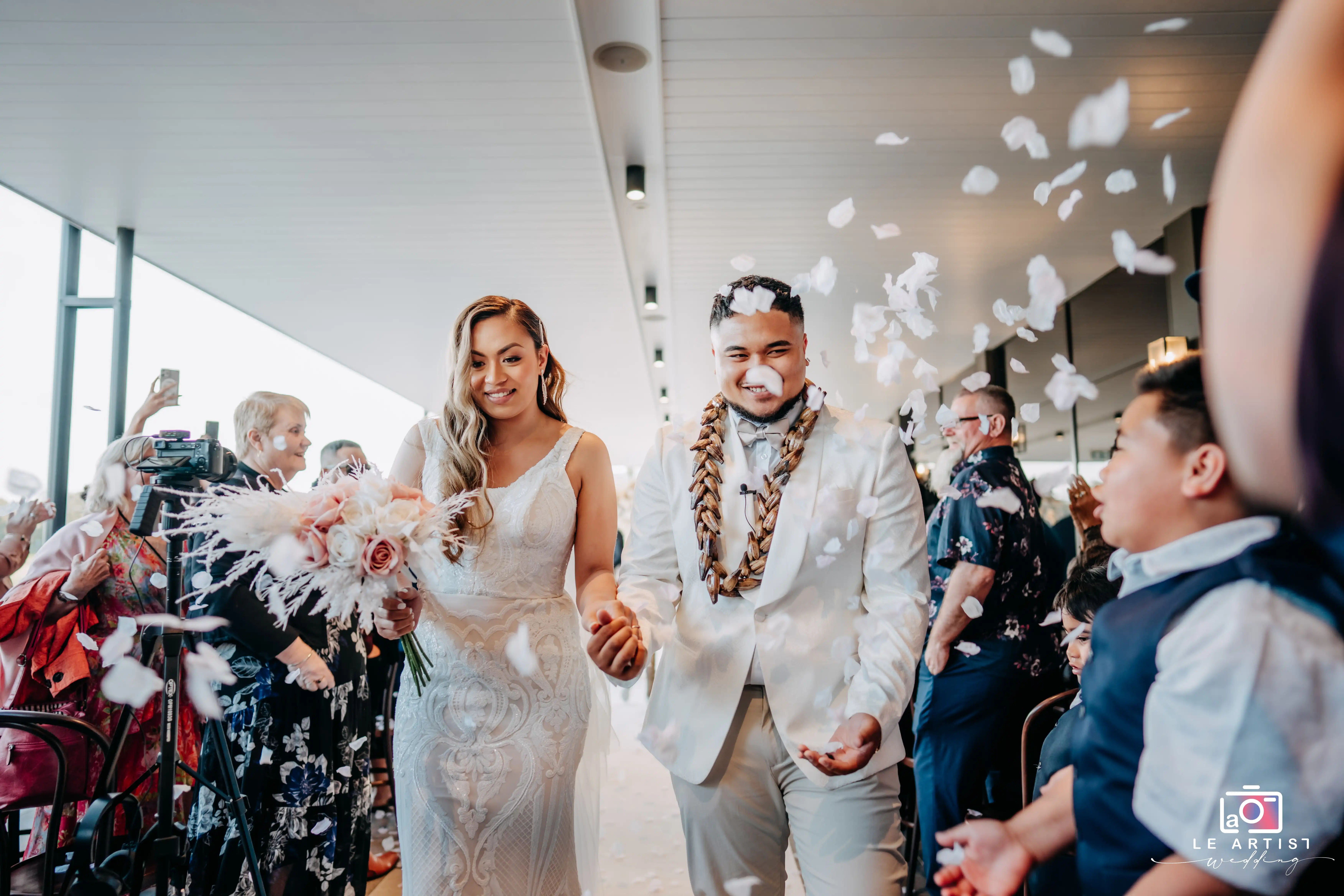 The newlyweds walk down the aisle, surrounded by flying petals and joyful guests capturing the moment. Le Artist Wedding