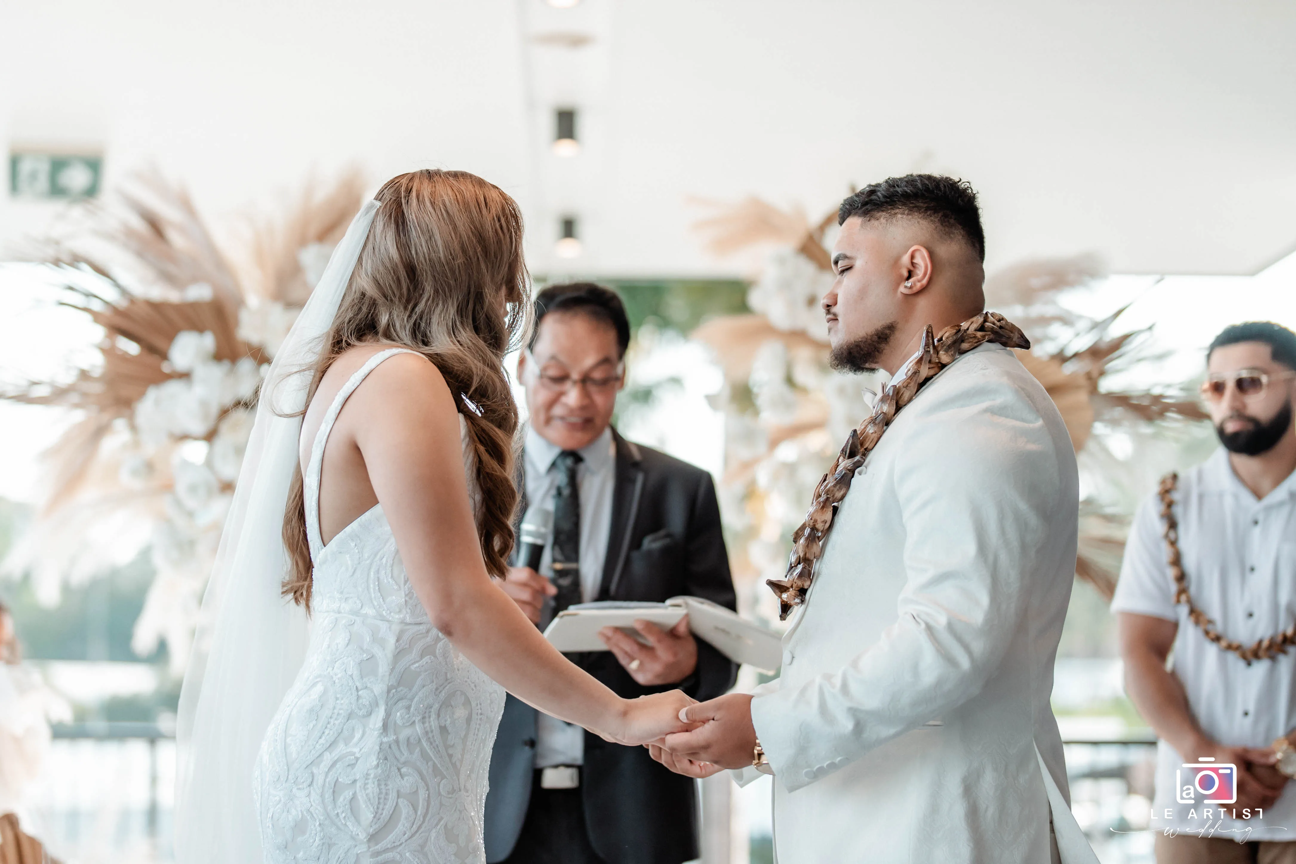 The couple holds hands as they exchange vows during their wedding ceremony, surrounded by elegant floral arrangements. Le Artist Wedding