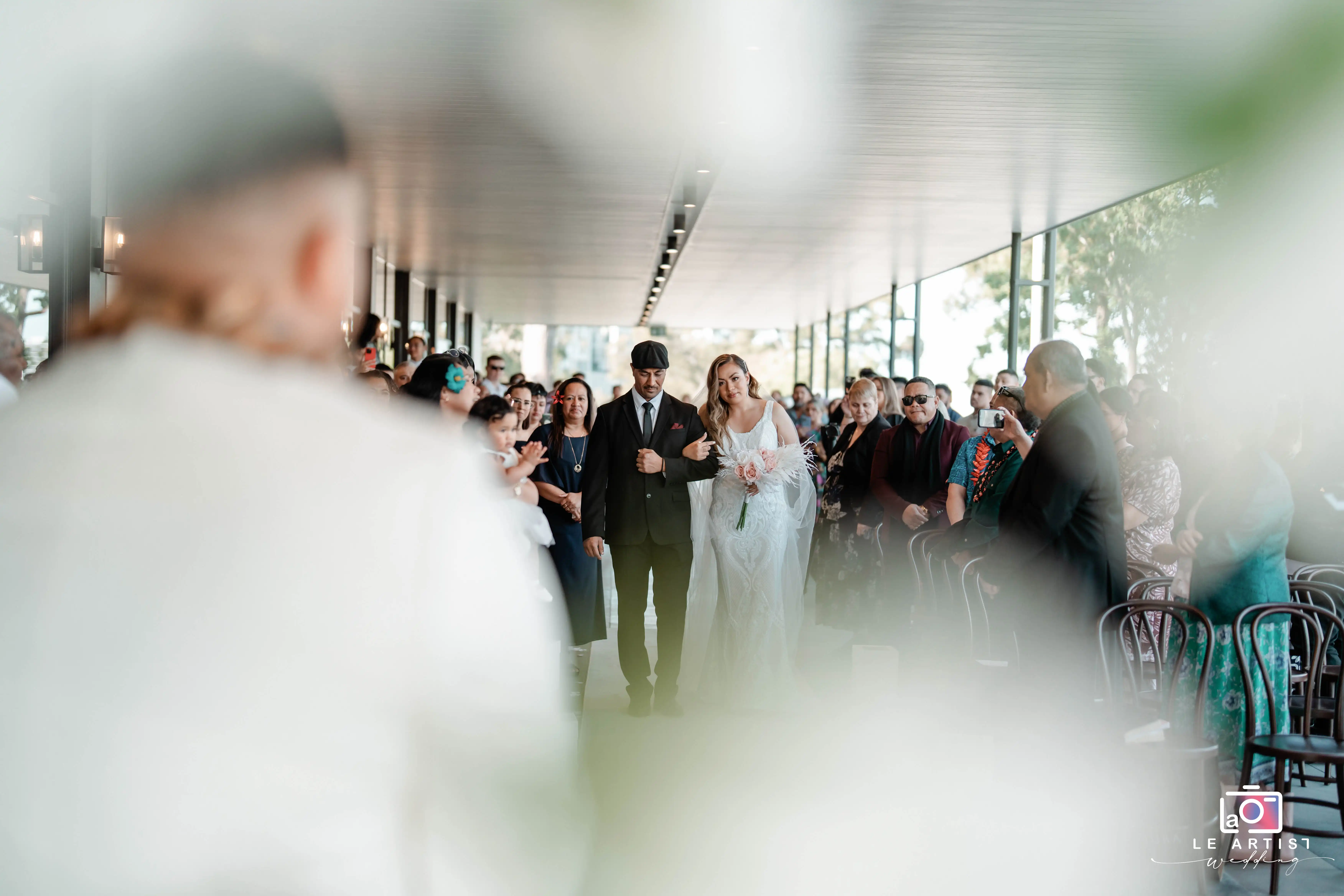 The bride walks down the aisle with her escort during the wedding ceremony, surrounded by joyful guests. Le Artist Wedding