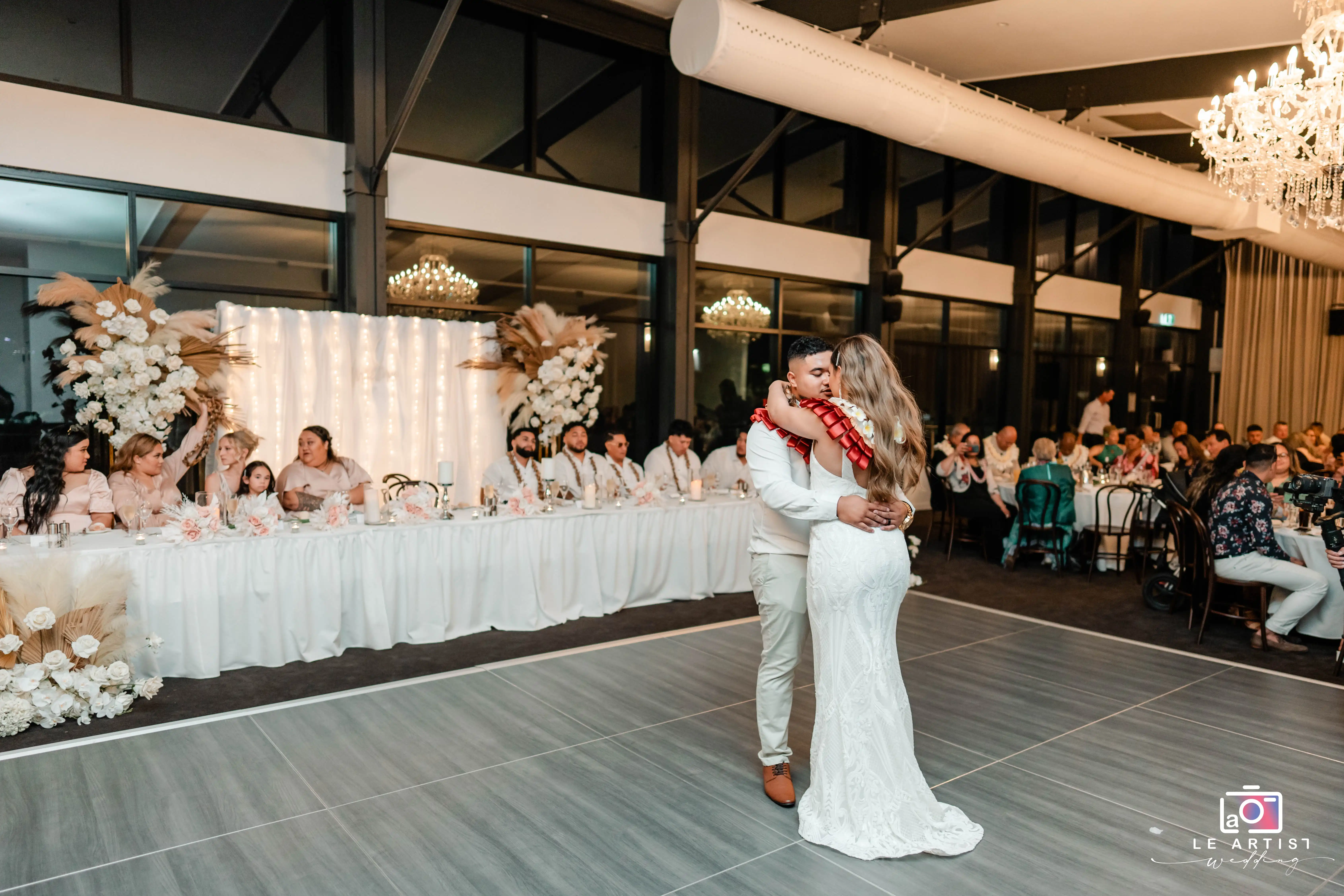 The couple shares a tender first dance at the wedding reception, with guests and the bridal party looking on warmly. Le Artist Wedding