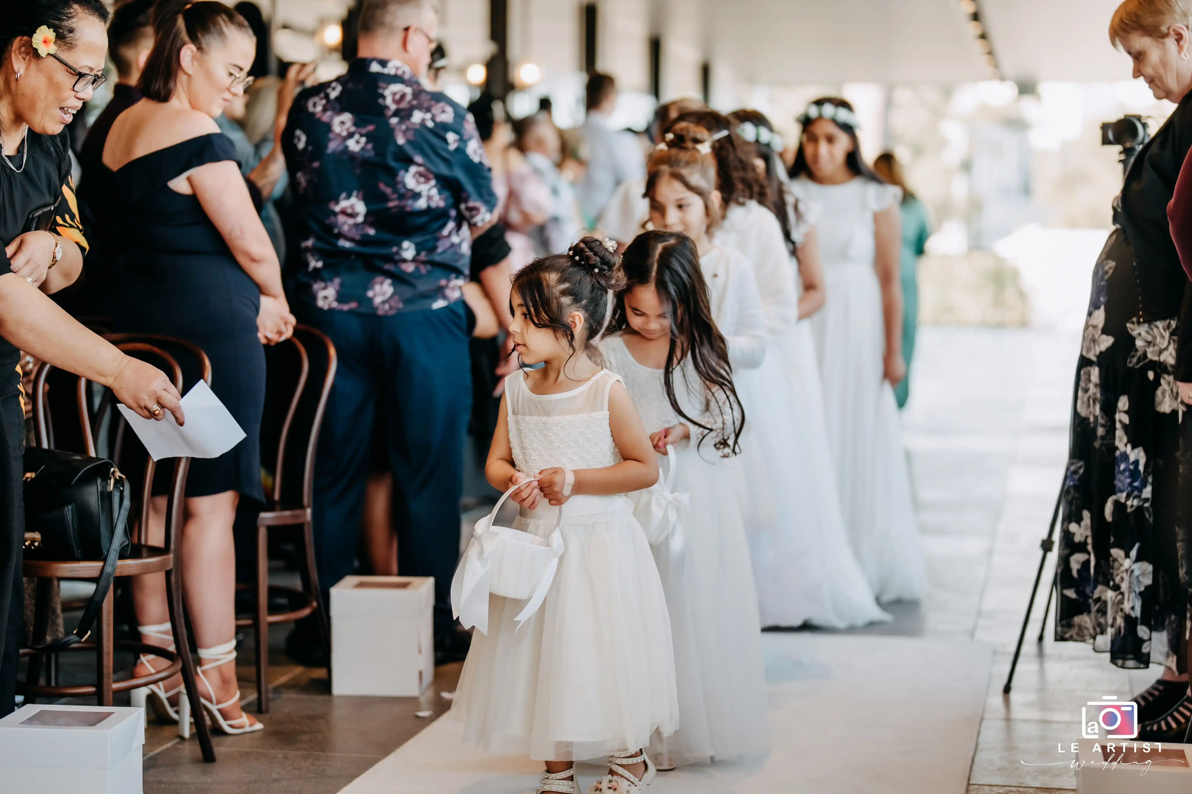 Flower girls lead the procession down the aisle, setting a charming scene as guests look on during the ceremony. Le Artist Wedding