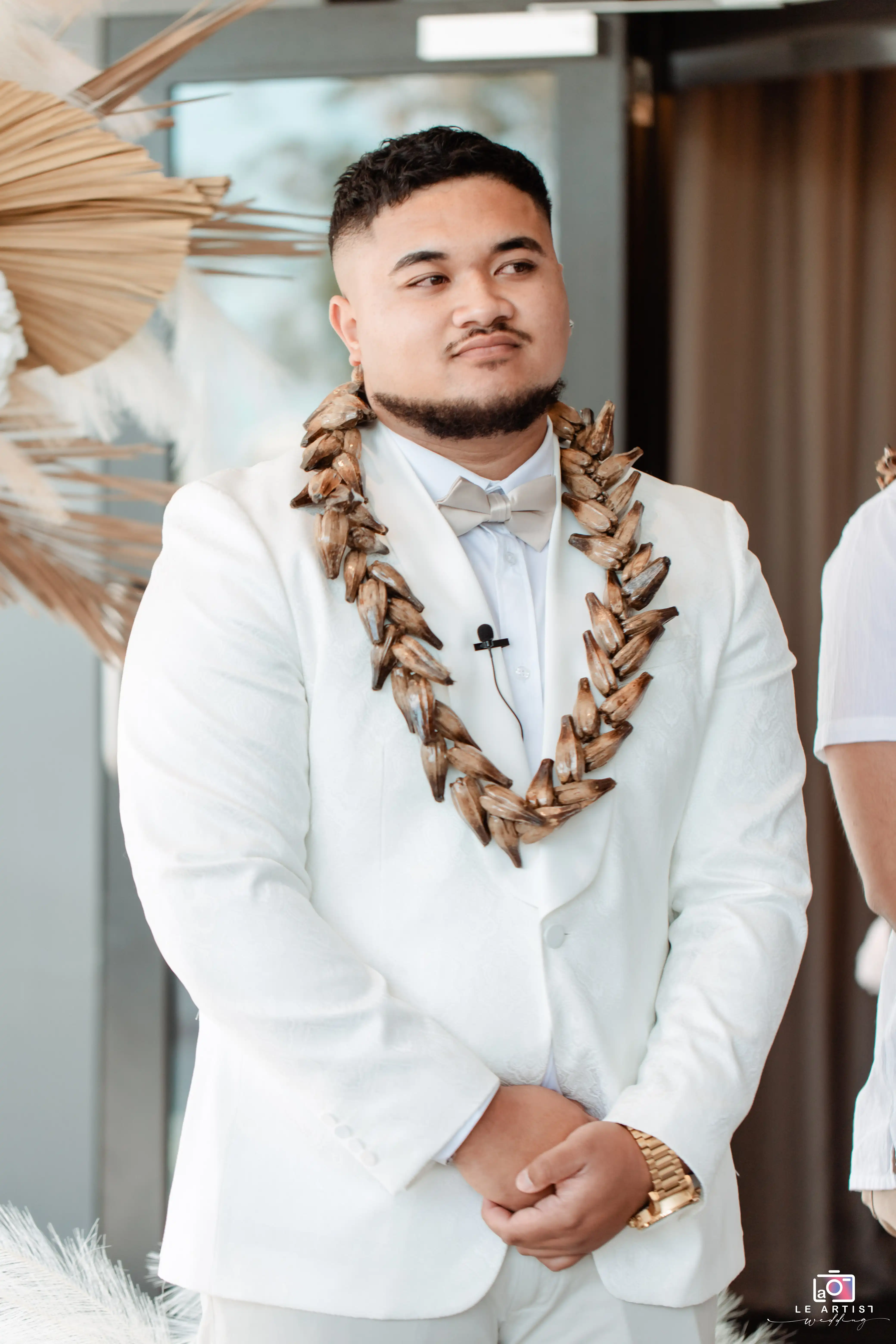 A groom stands with a solemn expression during the wedding ceremony, adorned in traditional attire and a unique lei. Le Artist Wedding