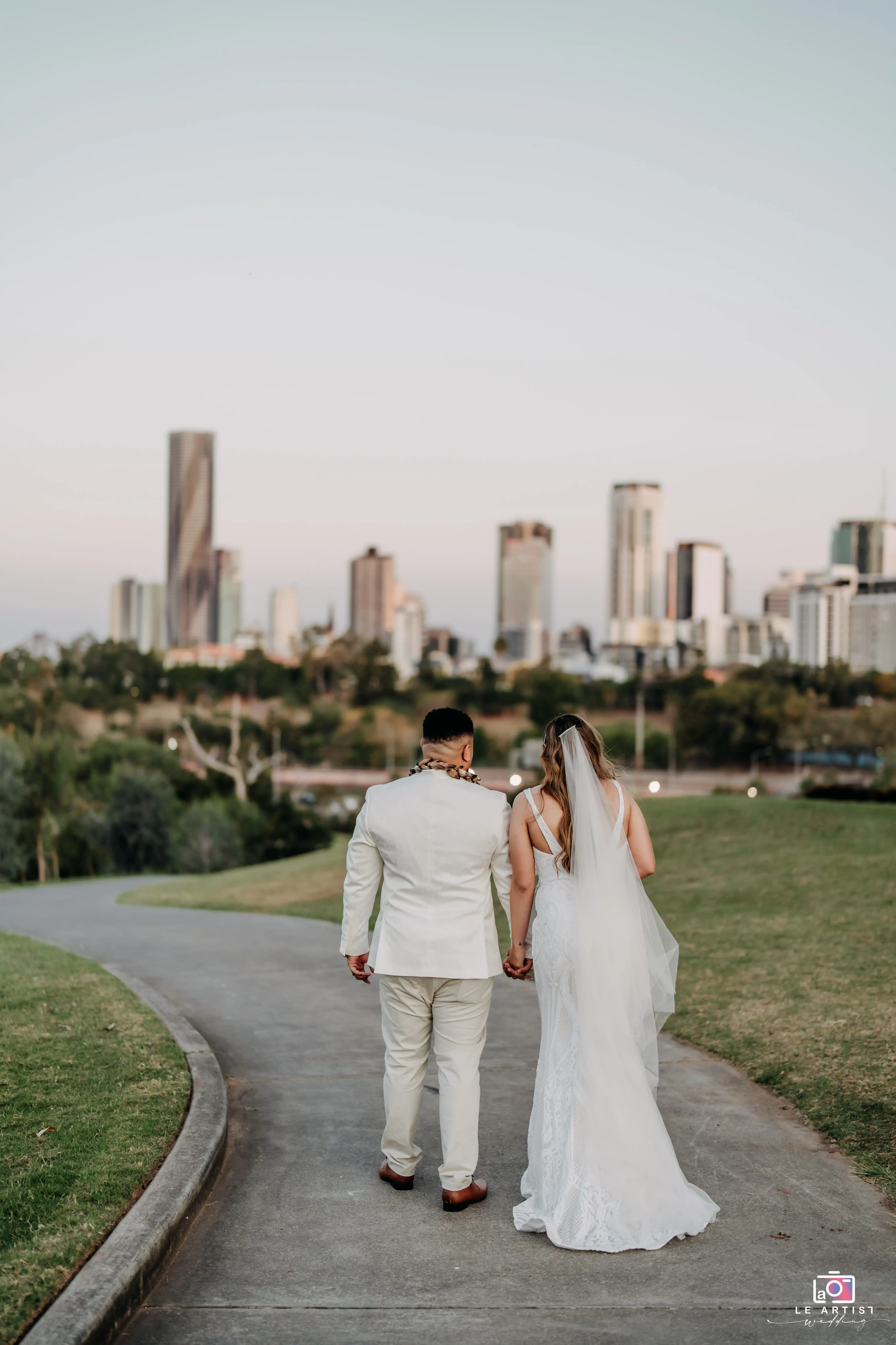 The newlyweds walk hand in hand, with a scenic city skyline in the background, capturing a serene moment on their special day. Le Artist Wedding