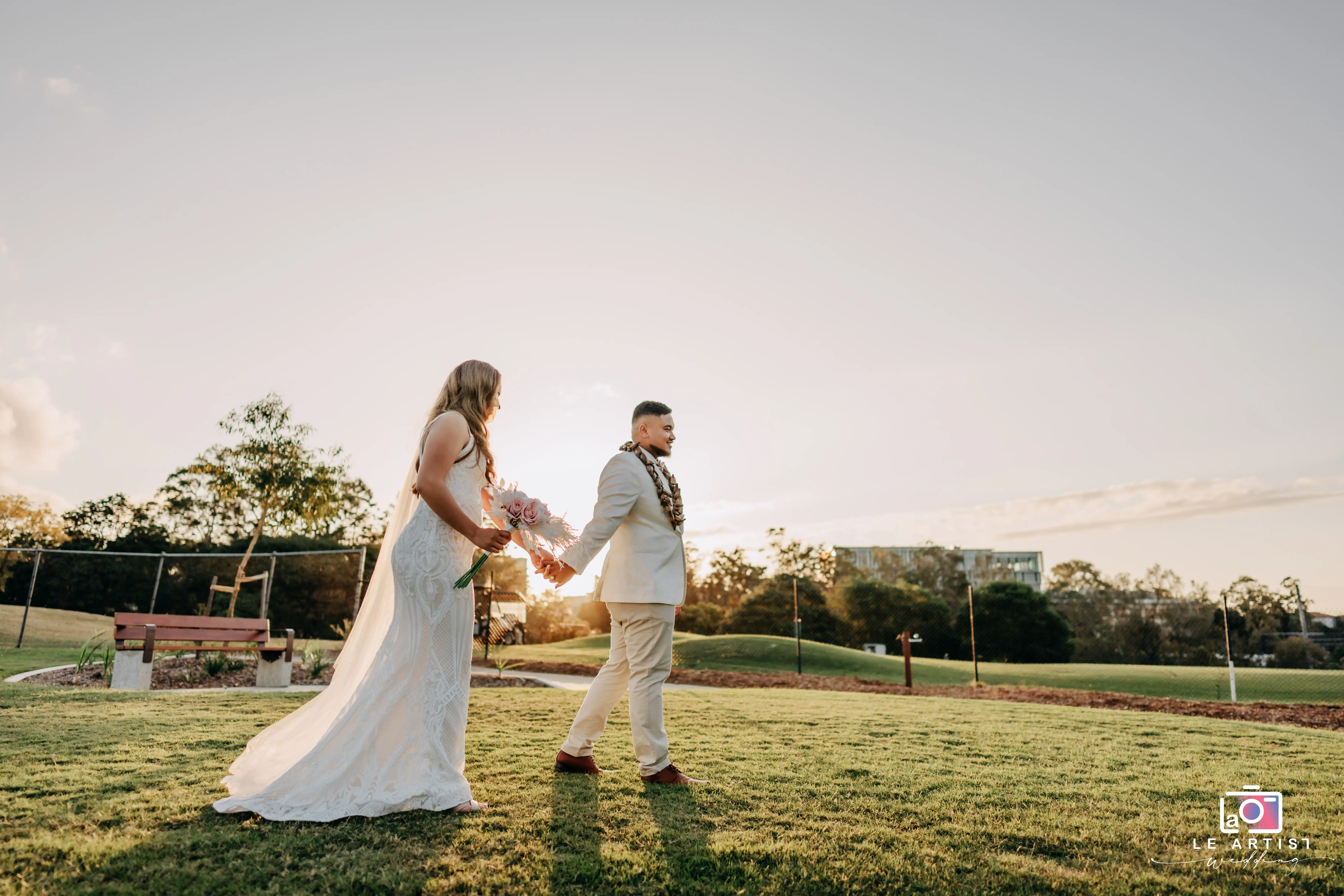 The newlyweds walk hand in hand across a sunlit field, capturing a serene moment together. Le Artist Wedding