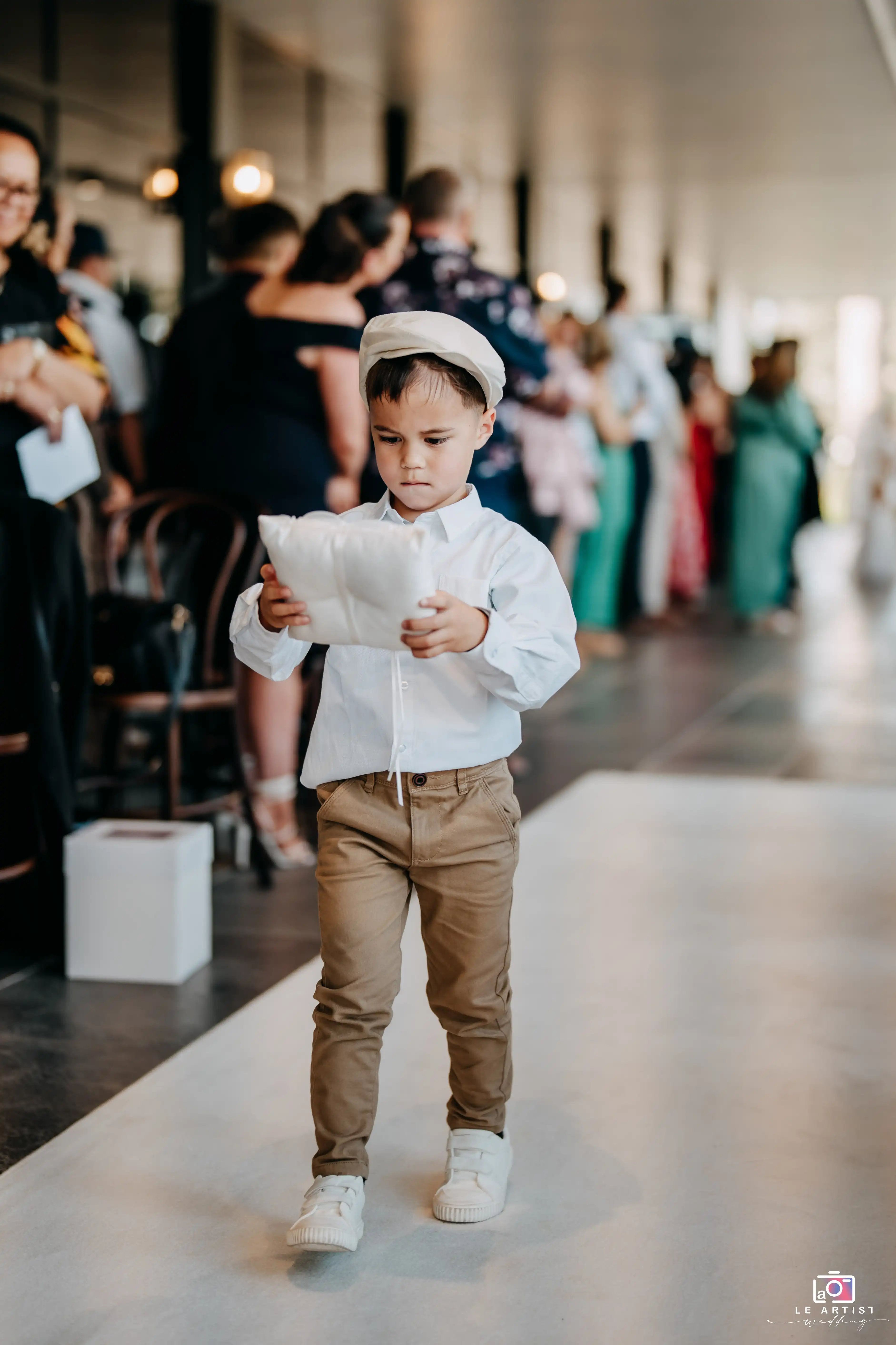 A young ring bearer walks solemnly down the aisle, clutching the pillow with rings as guests watch on. Le Artist Wedding