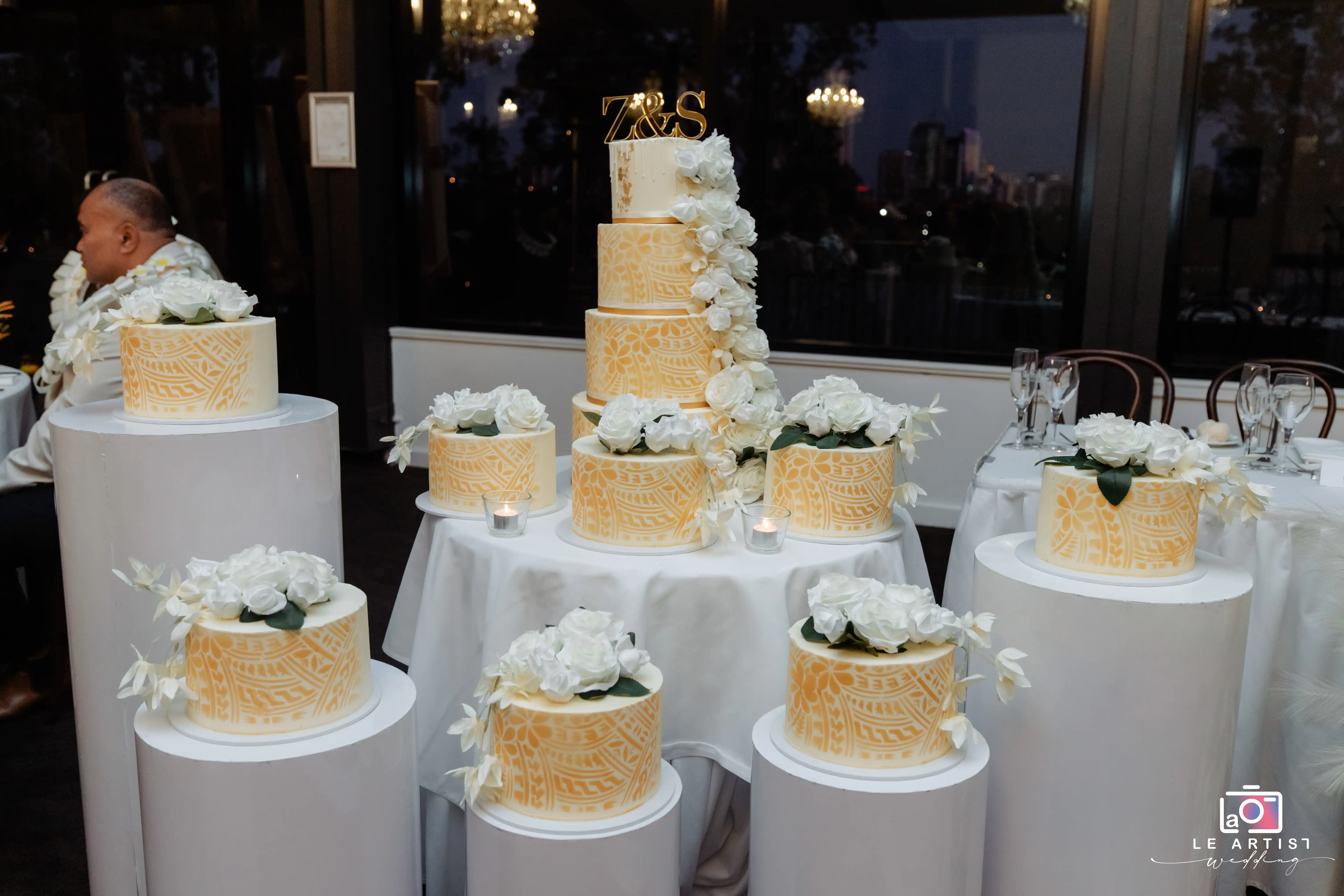 A beautifully decorated tiered wedding cake adorned with white roses and intricate designs, ready for the cake cutting ceremony. Le Artist Wedding