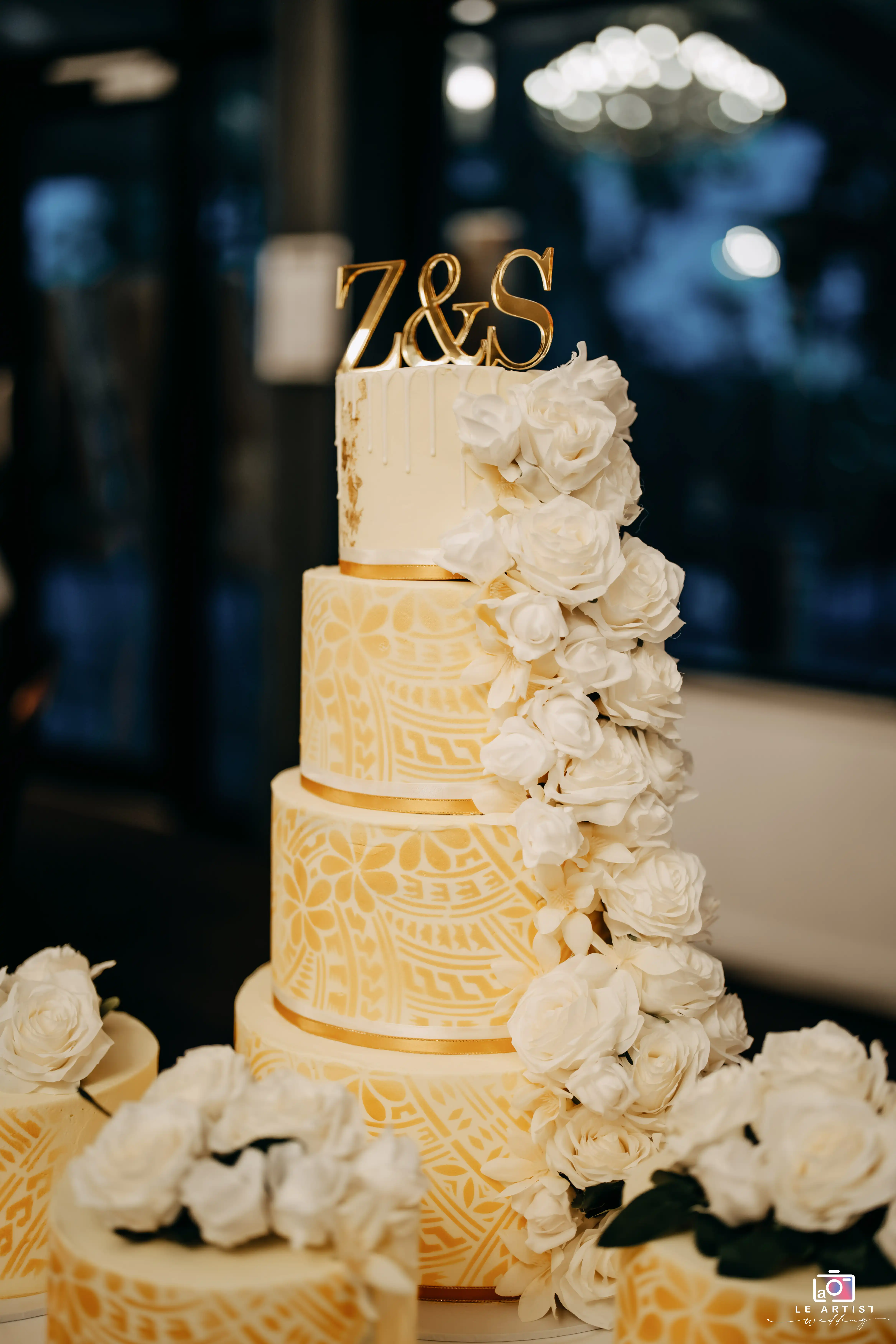 A beautifully decorated wedding cake adorned with intricate designs and white roses, topped with initials Z & S, ready for the cake cutting ceremony. Le Artist Wedding