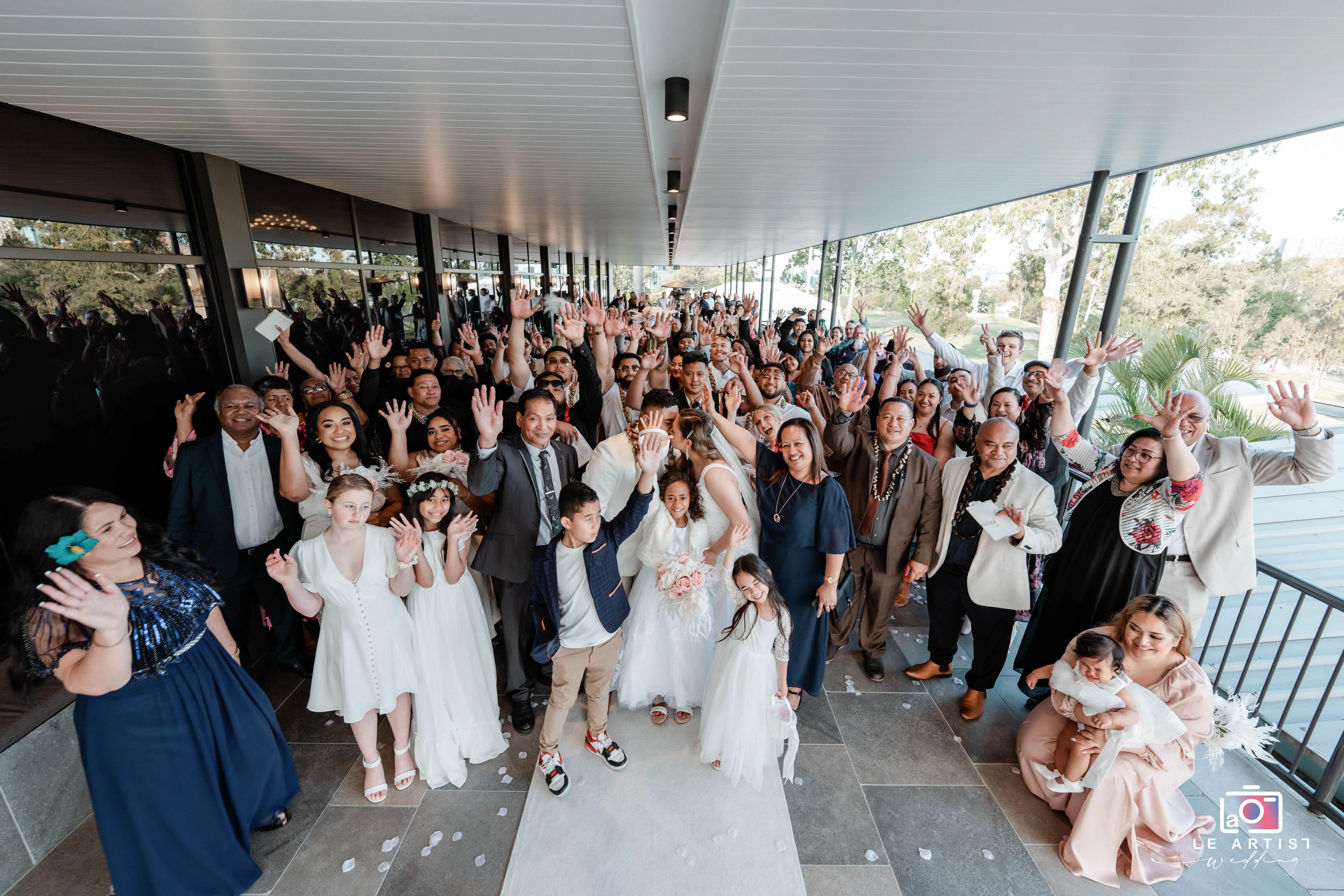 A joyful gathering of wedding guests celebrates as the couple makes their iconic processional walk, with smiles and raised hands in a corridor adorned with natural light. Le Artist Wedding
