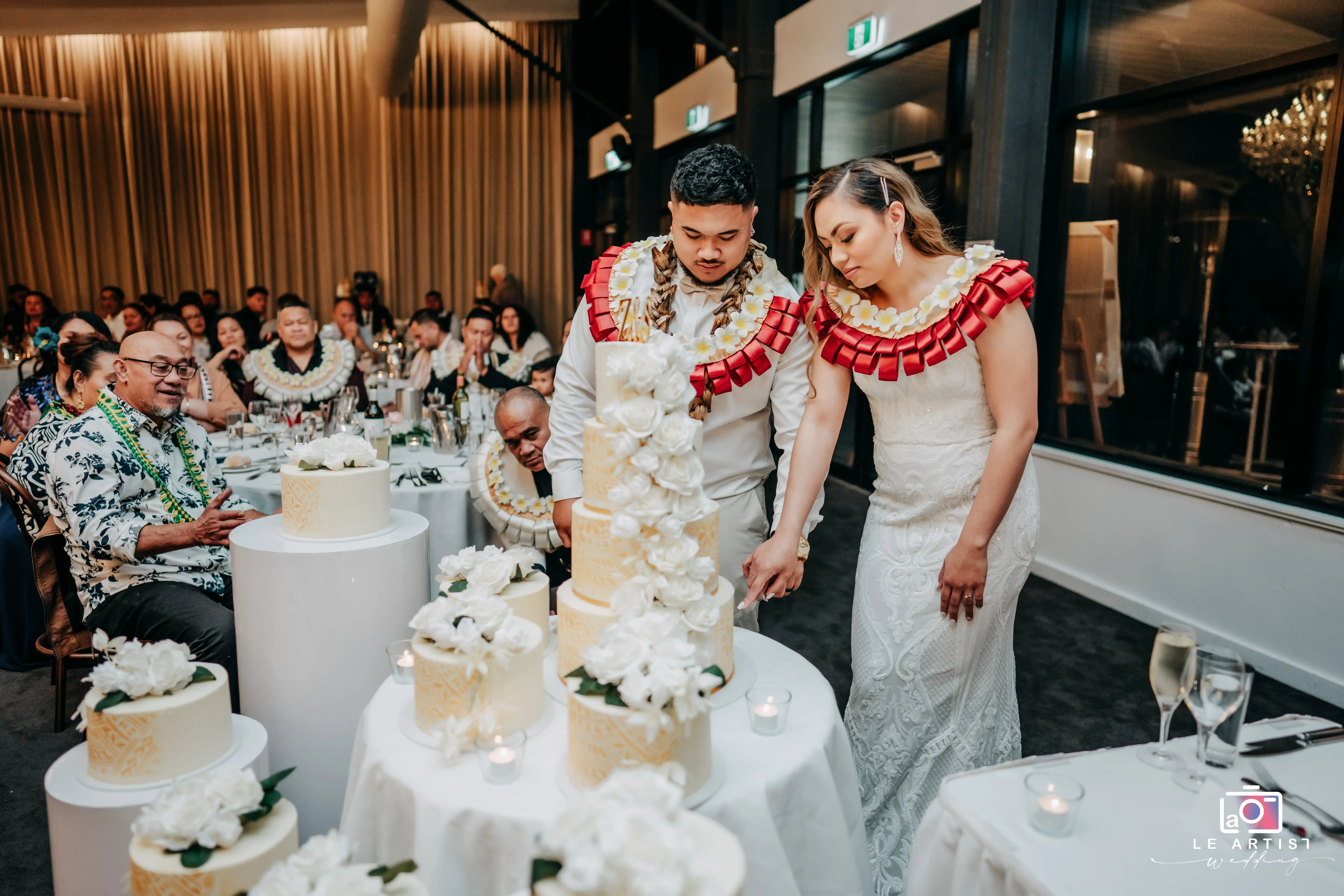 The couple shares a moment as they cut the wedding cake, adorned with white flowers, while guests look on in celebration. Le Artist Wedding