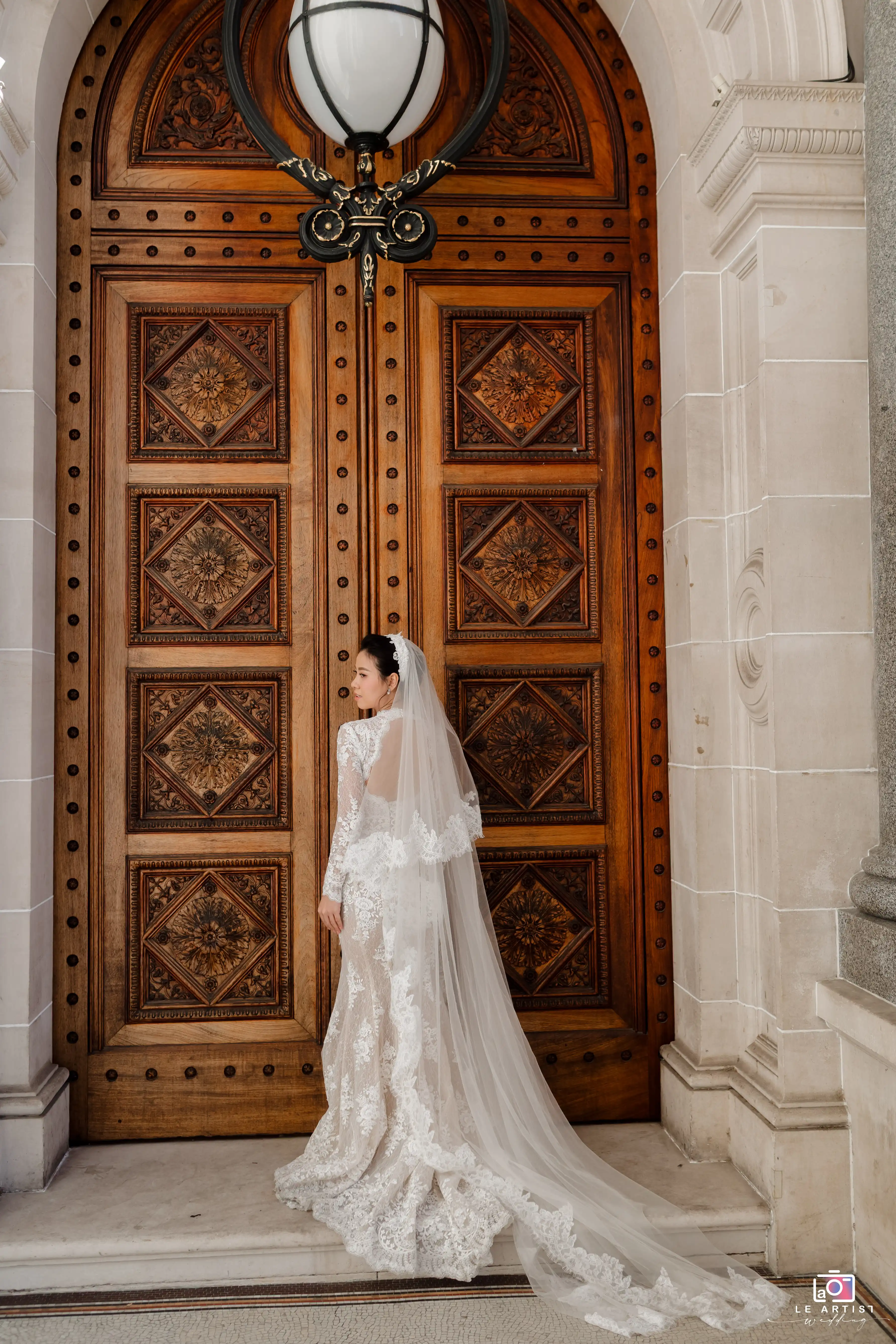 A stunning bridal portrait featuring the bride in an exquisite lace gown with a flowing veil, standing elegantly against an intricately carved wooden door. Le Artist Wedding