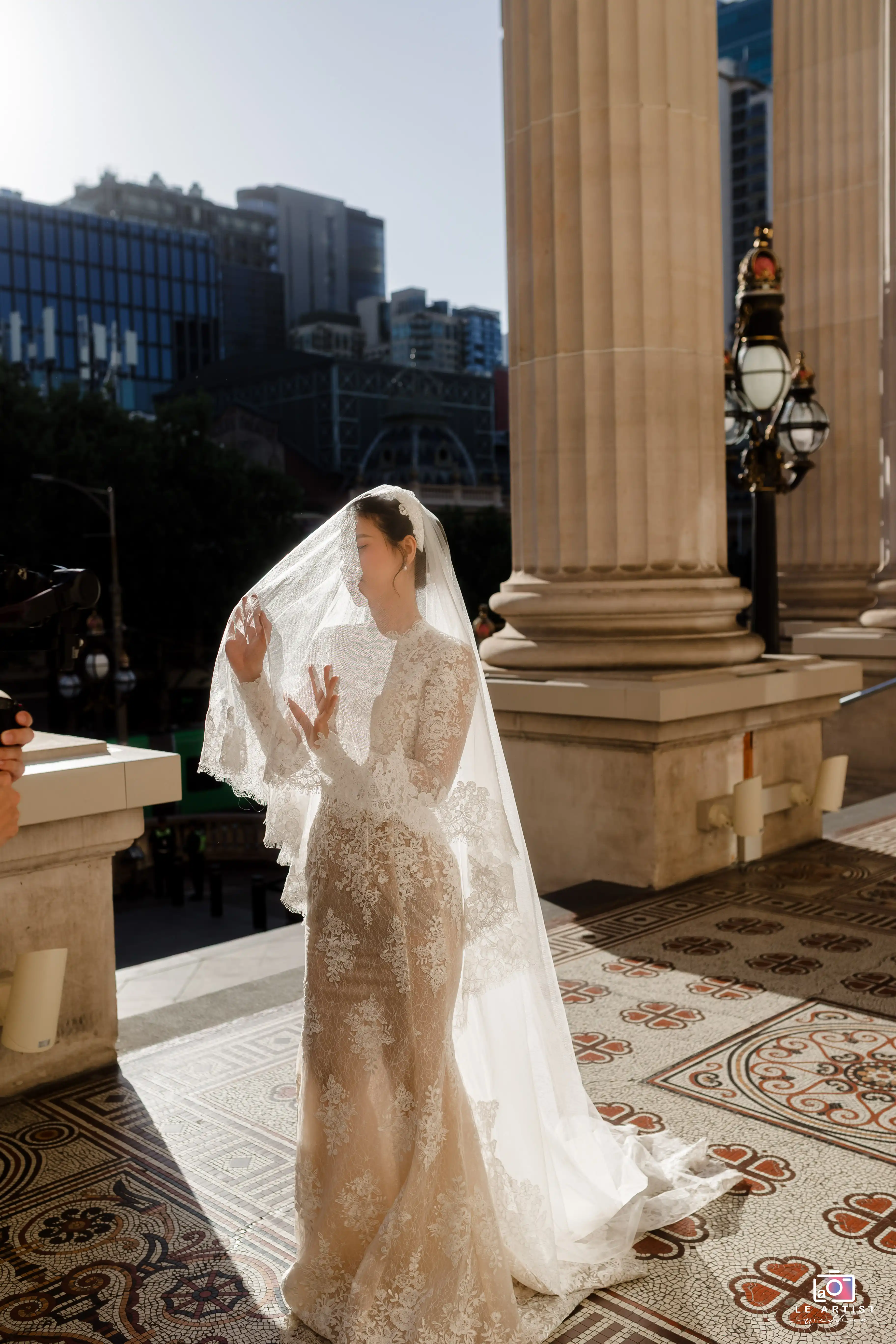 The bride stands gracefully on the steps, her lace dress glimmering in the sunlight as she adjusts her veil. The cityscape forms a unique backdrop, blending urban architecture with the classic elegance of the wedding. Le Artist Wedding