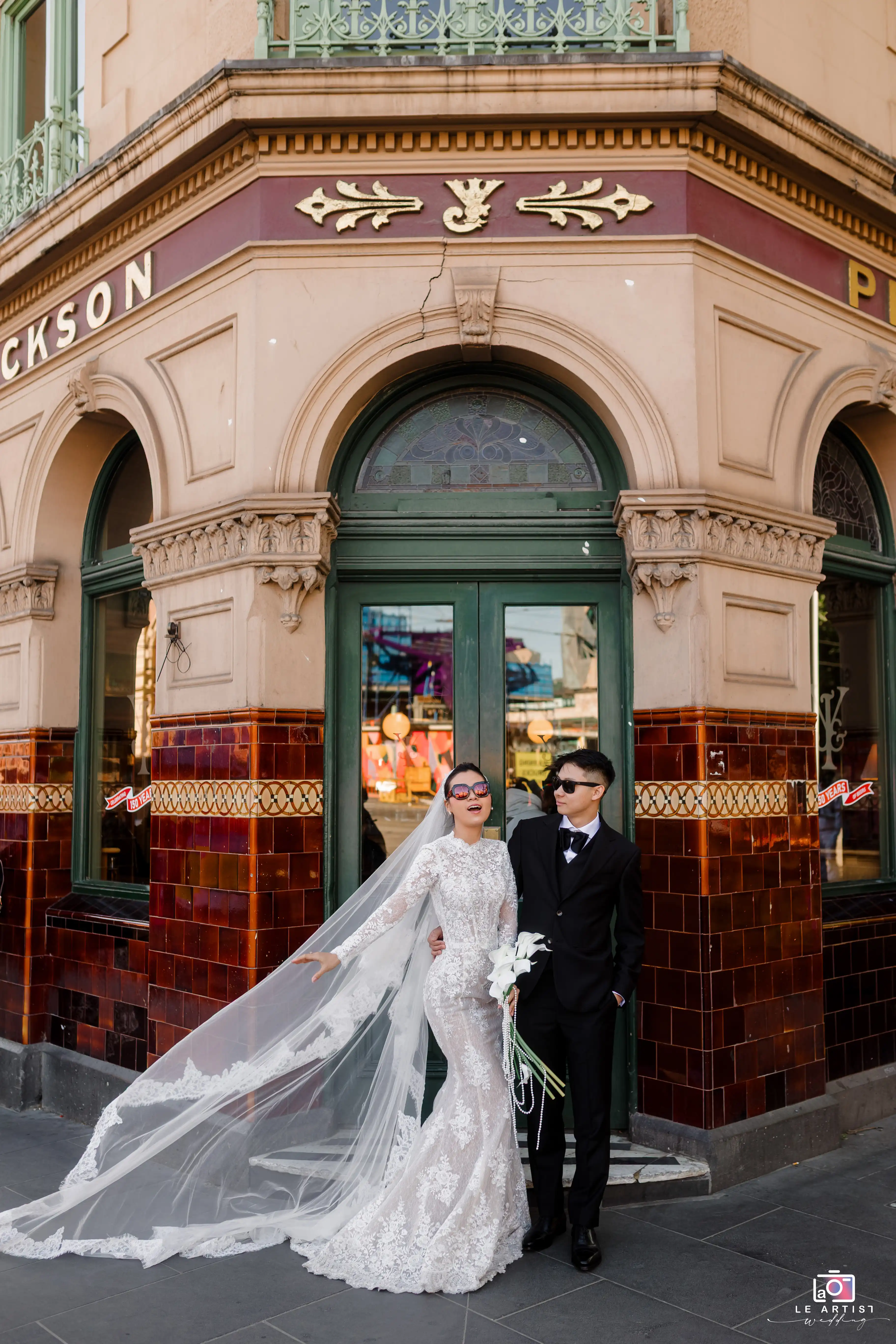 The bride and groom share a joyful moment outside a beautifully ornate building, exuding elegance with their sunglasses and stylish attire. Le Artist Wedding