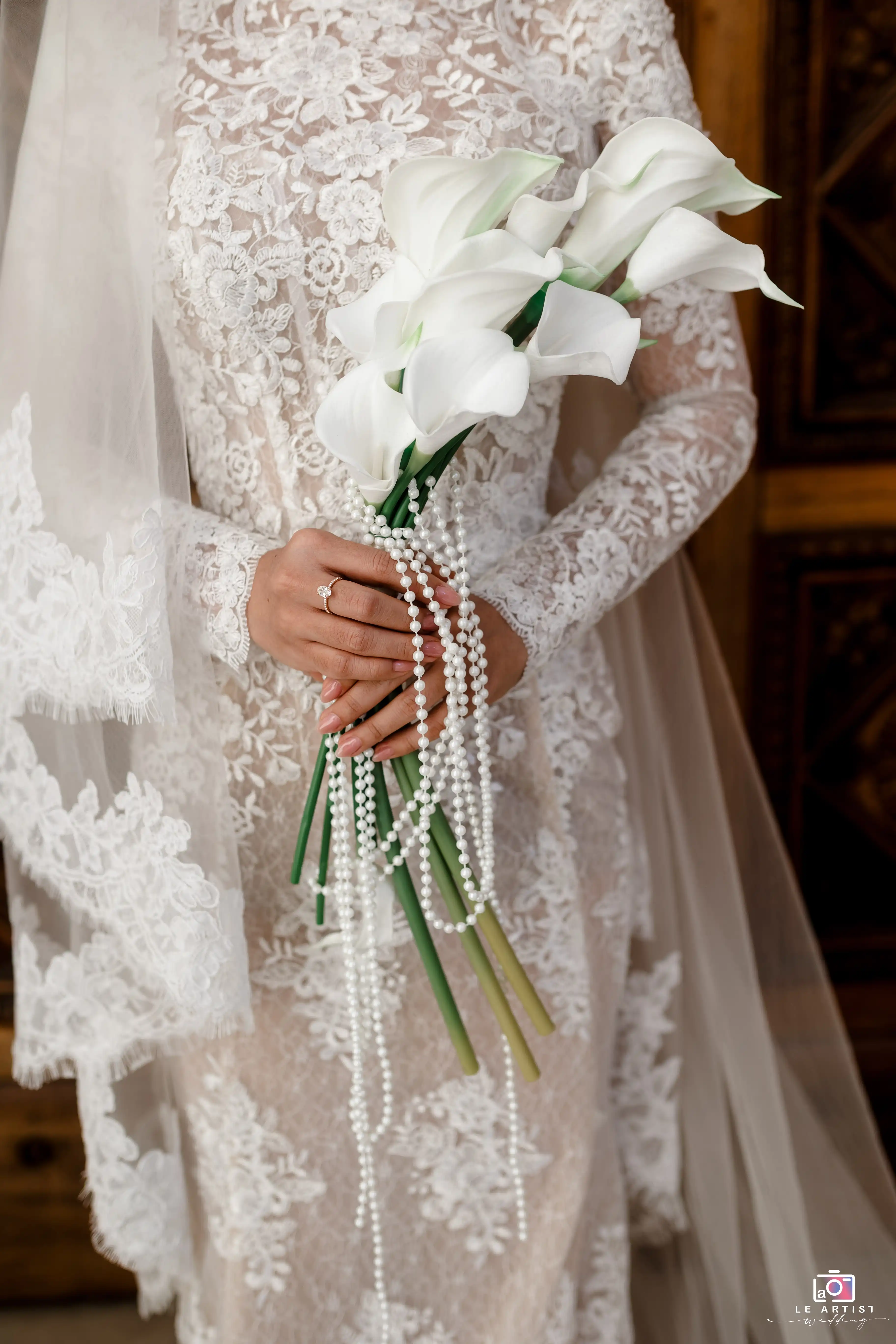 Bride holding a bouquet of elegant white calla lilies adorned with delicate pearl strings, showcasing intricate lace details of her wedding dress. Le Artist Wedding