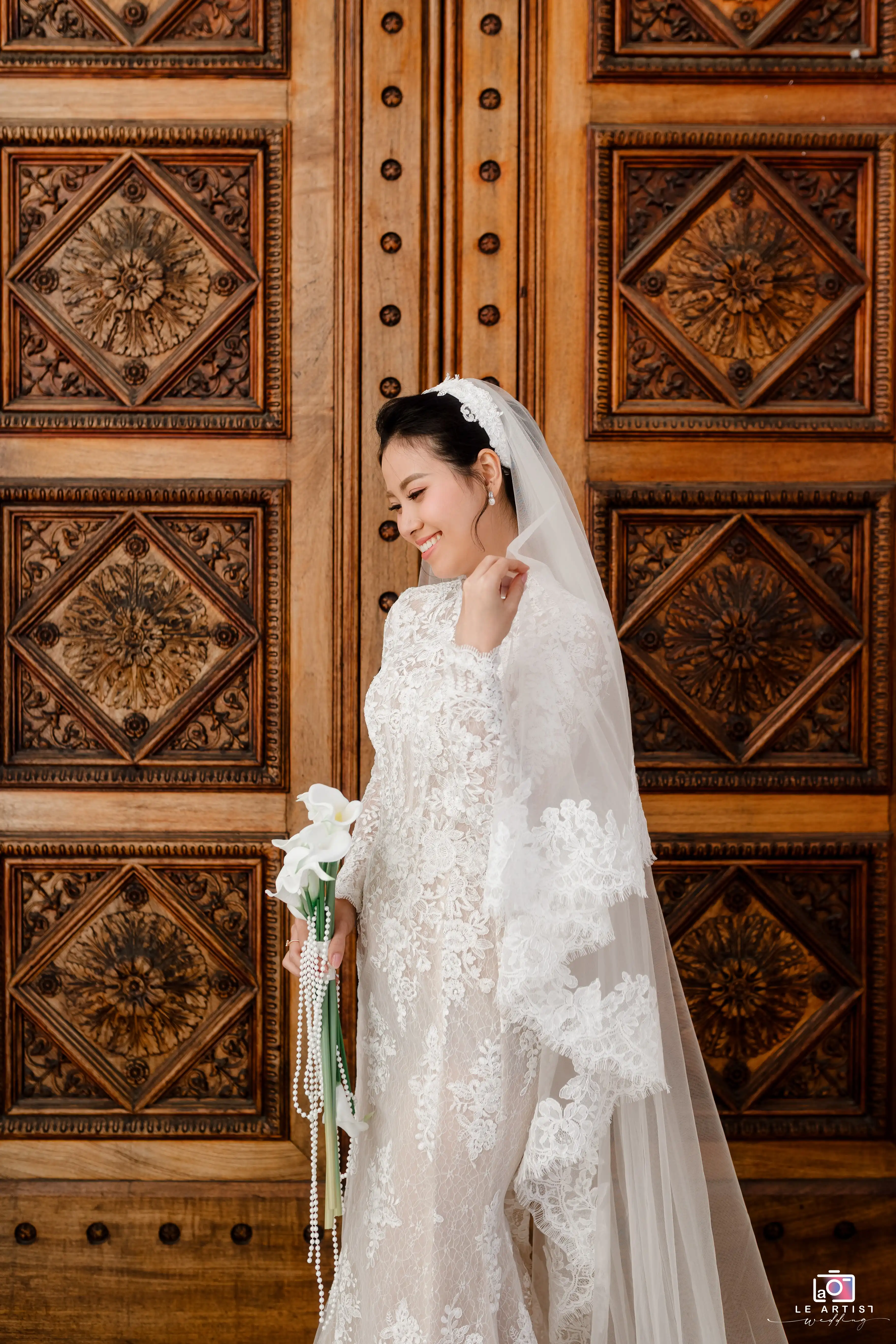 The bride stands gracefully in an intricately designed lace gown, holding a delicate bouquet of white lilies, against a beautifully carved wooden backdrop. Le Artist Wedding