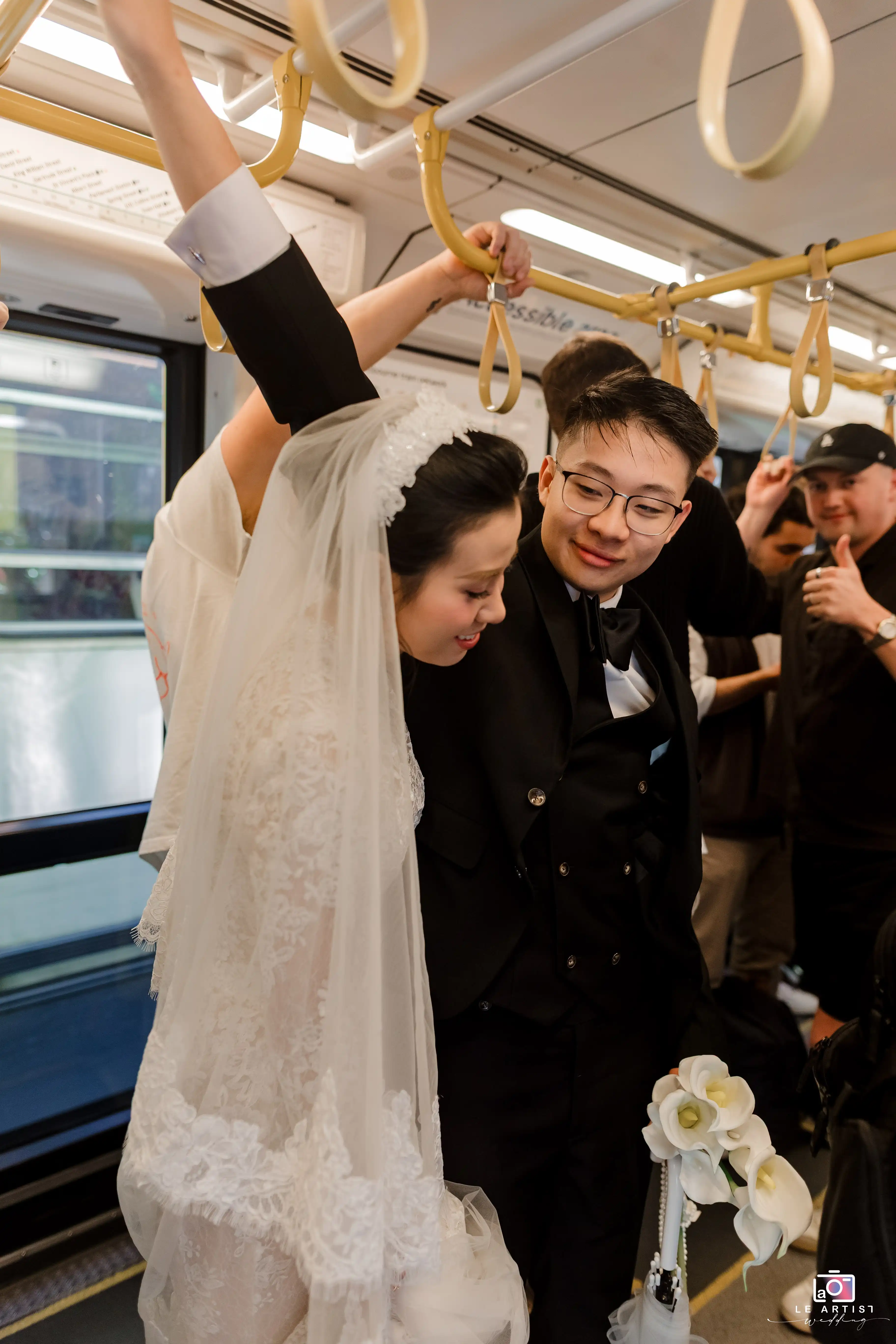 A joyful bride and groom share a tender moment on public transport, with the bride in her elegant gown and veil, and the groom in a suit, surrounded by onlookers. Le Artist Wedding