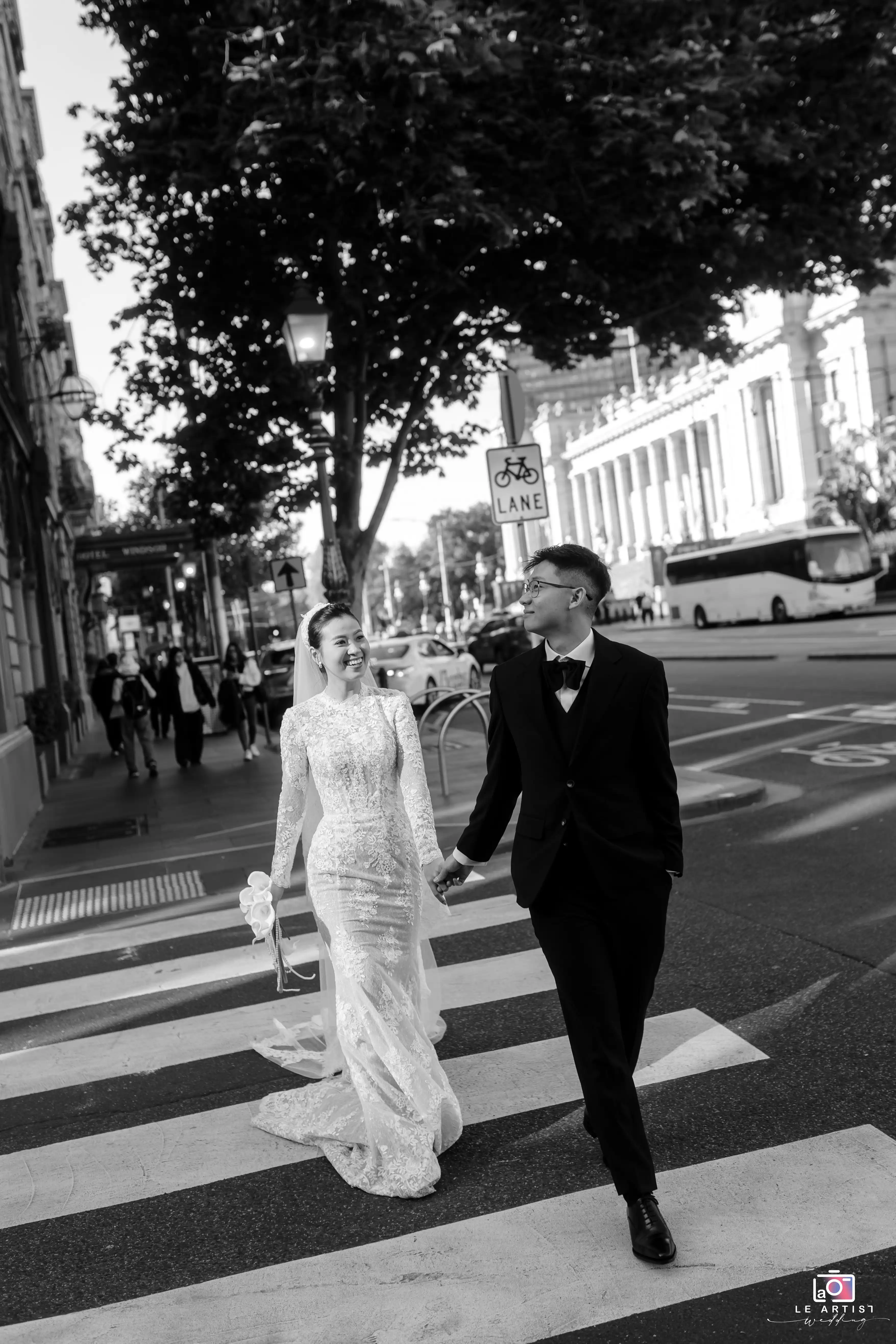The bride and groom share a joyous moment together as they walk hand in hand across a city street, their happiness evident in their smiles. Le Artist Wedding