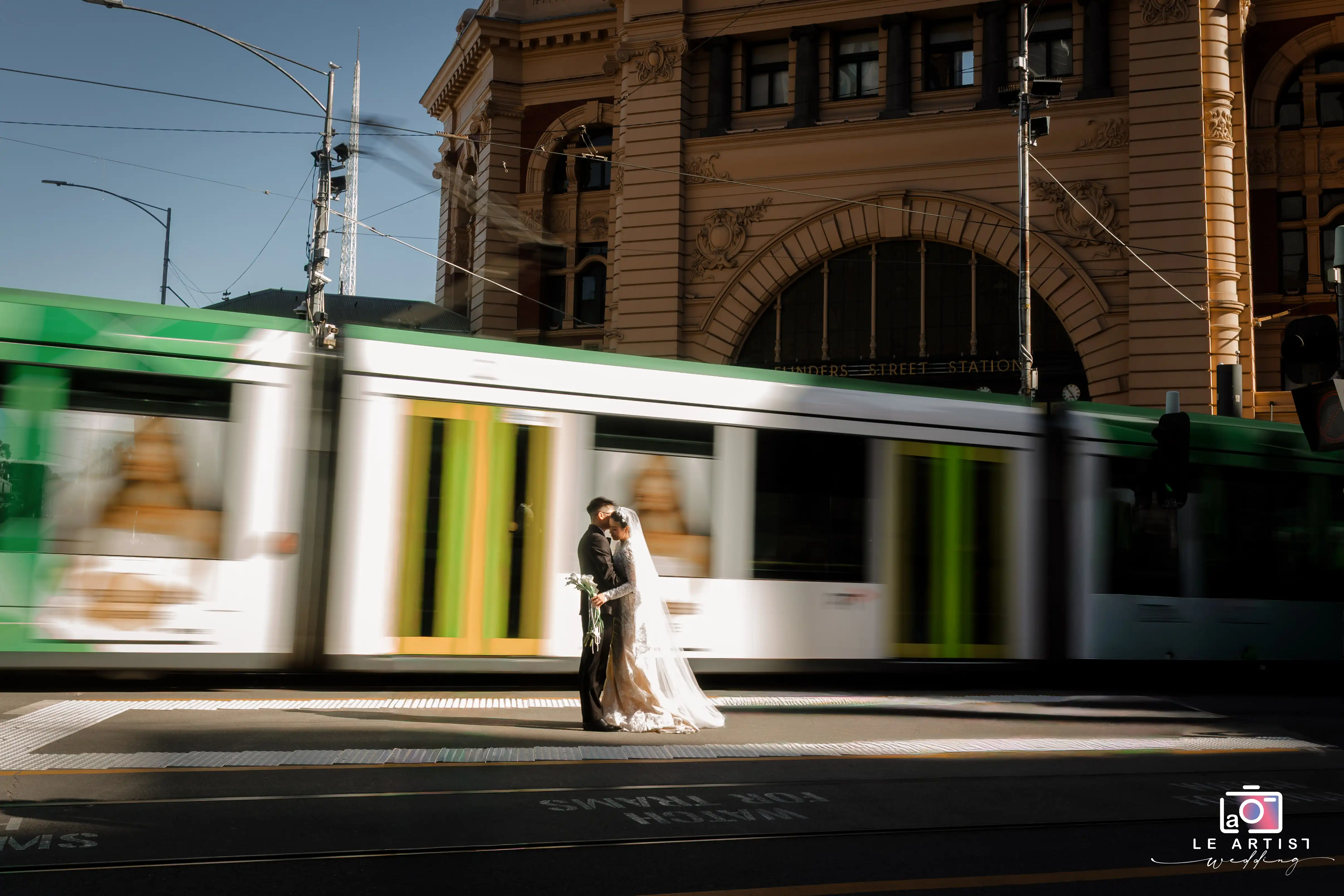 A couple shares a tender moment in front of a bustling cityscape with a tram speeding past, symbolizing love amidst the hustle and bustle. Le Artist Wedding
