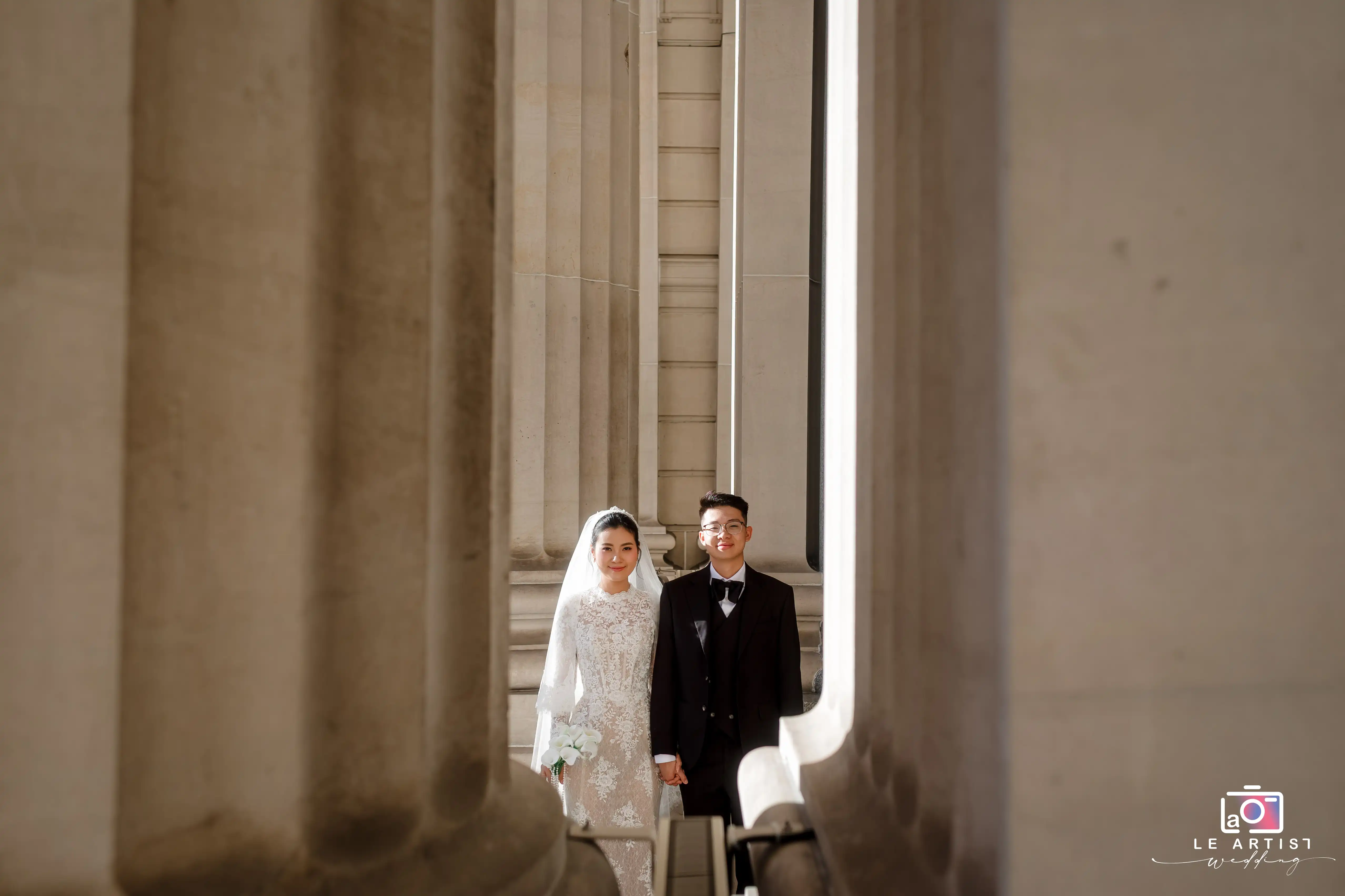 A radiant couple stands between grand architectural columns, capturing a timeless wedding moment. Le Artist Wedding