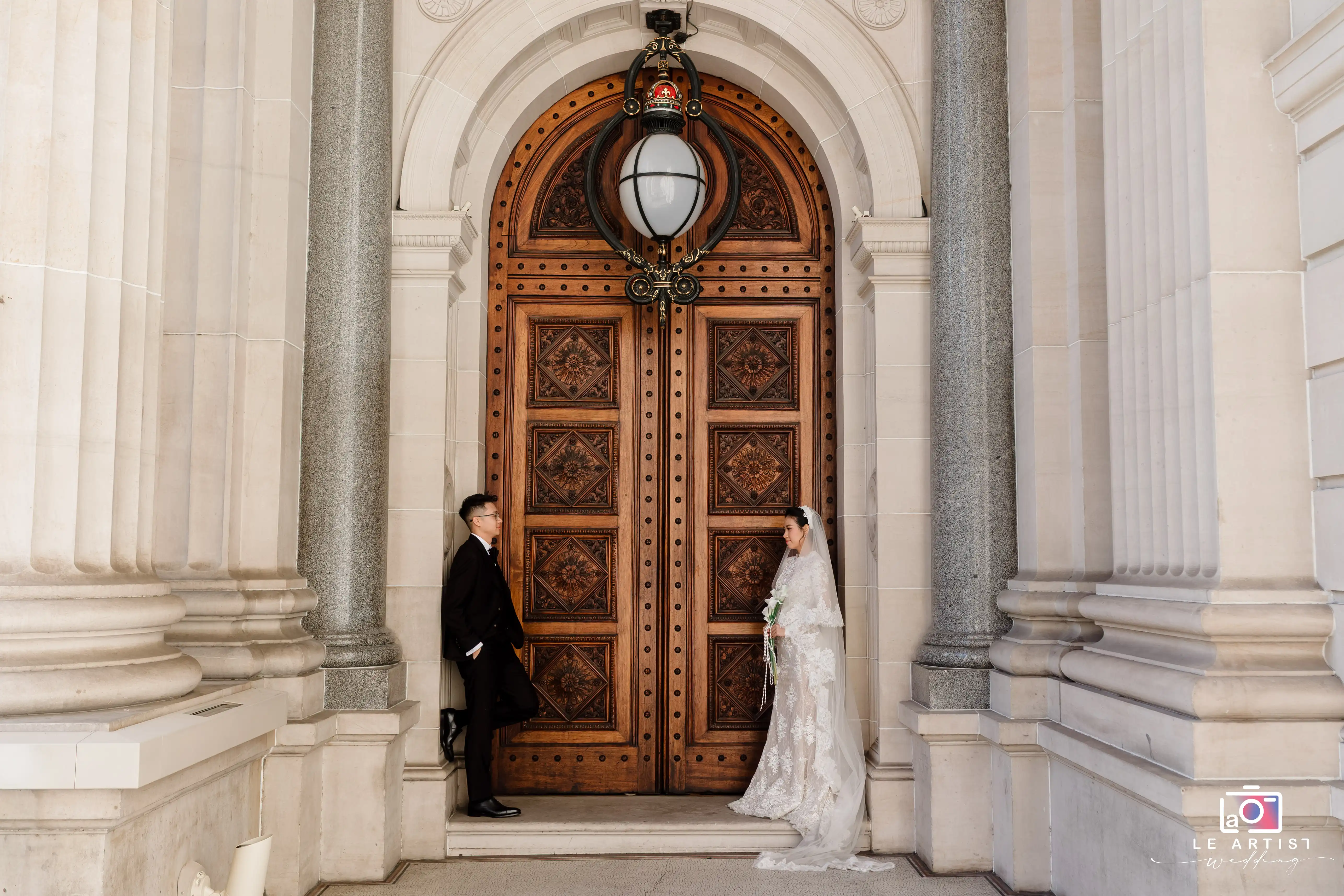 A couple shares a quiet moment outside an ornate wooden doorway, set against a backdrop of classical architecture. Le Artist Wedding