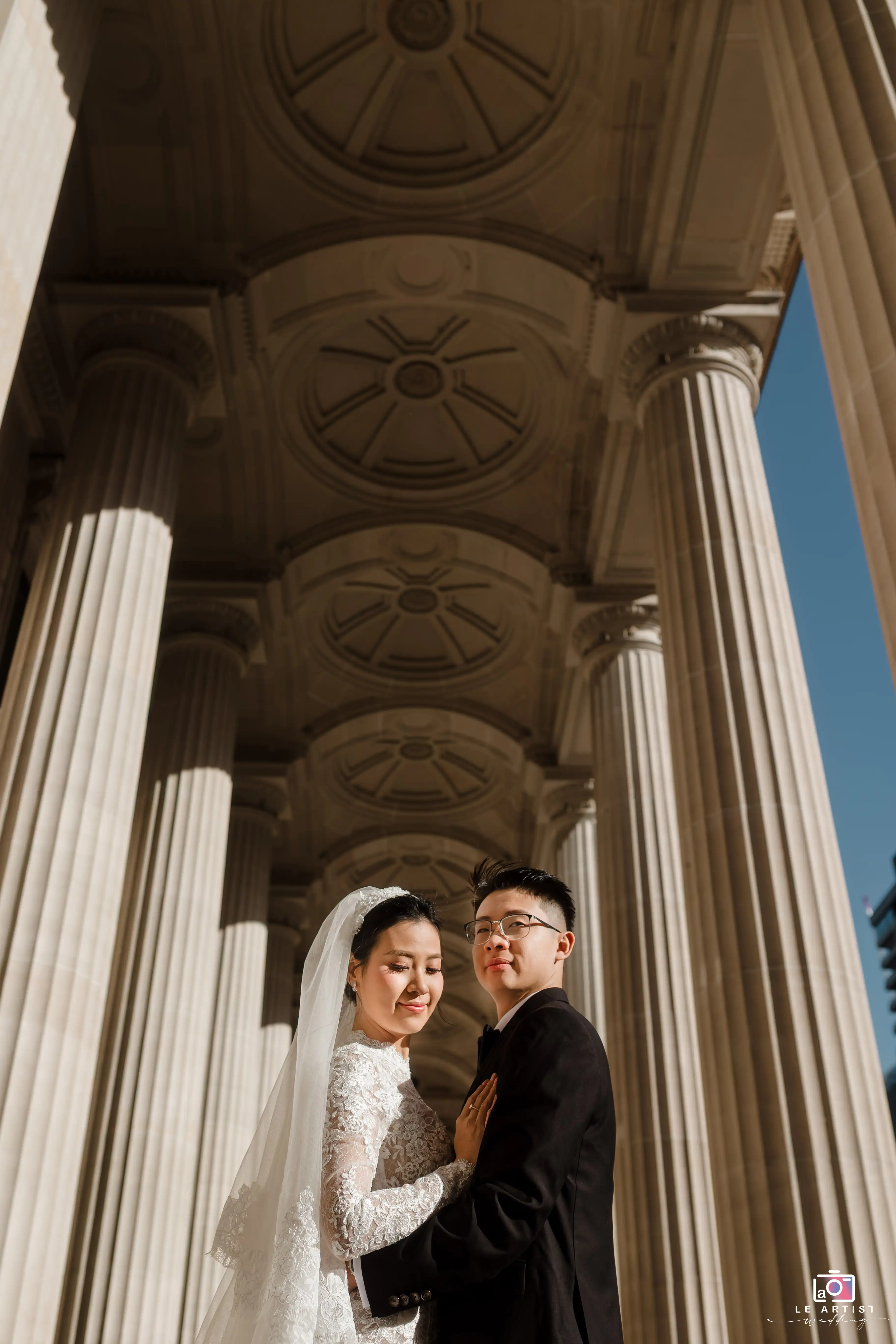 The couple stands gracefully under grand architectural columns, bathed in warm sunlight, capturing a timeless moment of elegance and love. Le Artist Wedding