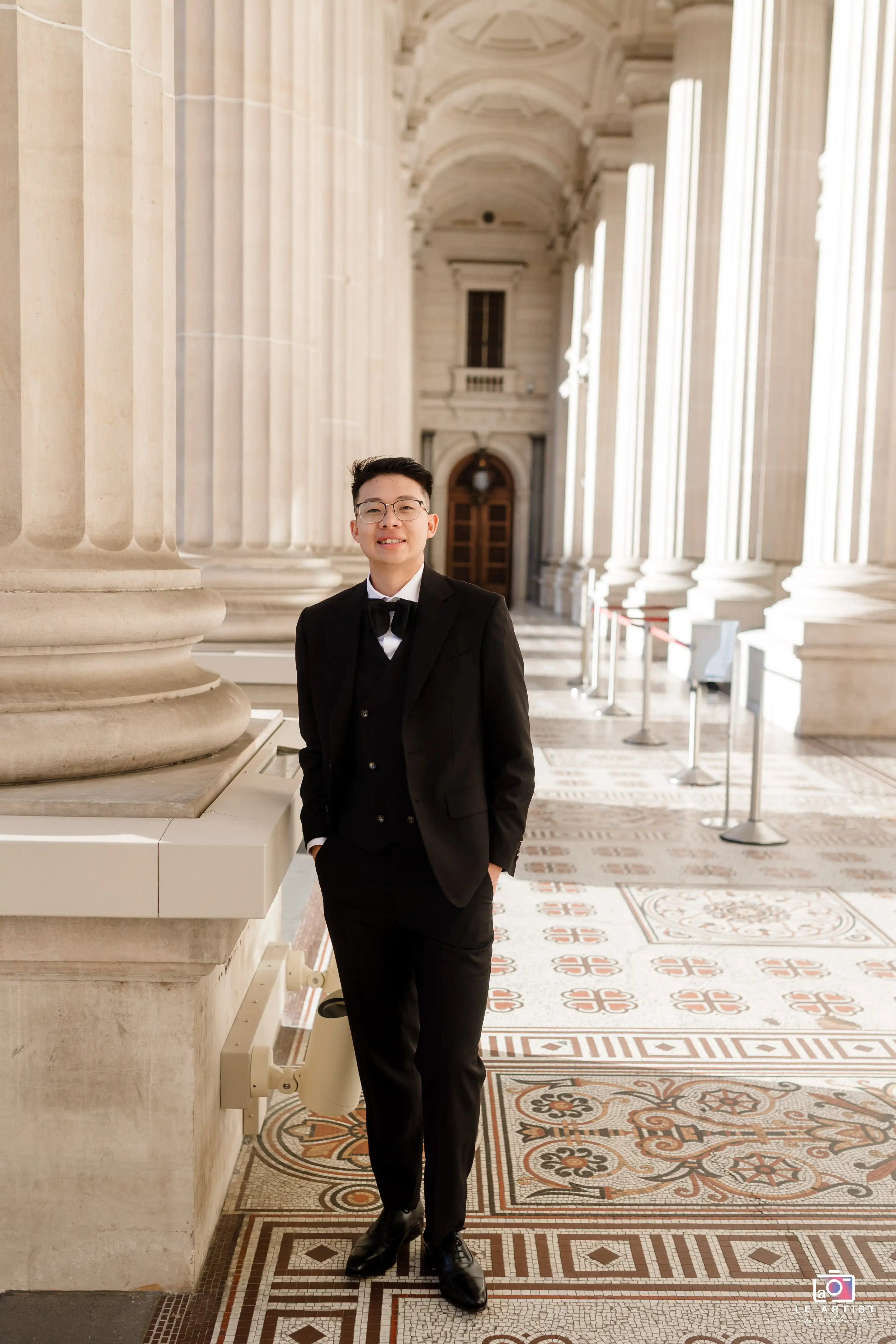 A groom poses elegantly in a classic black suit, standing amidst grand architectural columns and intricate tiled flooring, capturing a sophisticated moment before the wedding. Le Artist Wedding