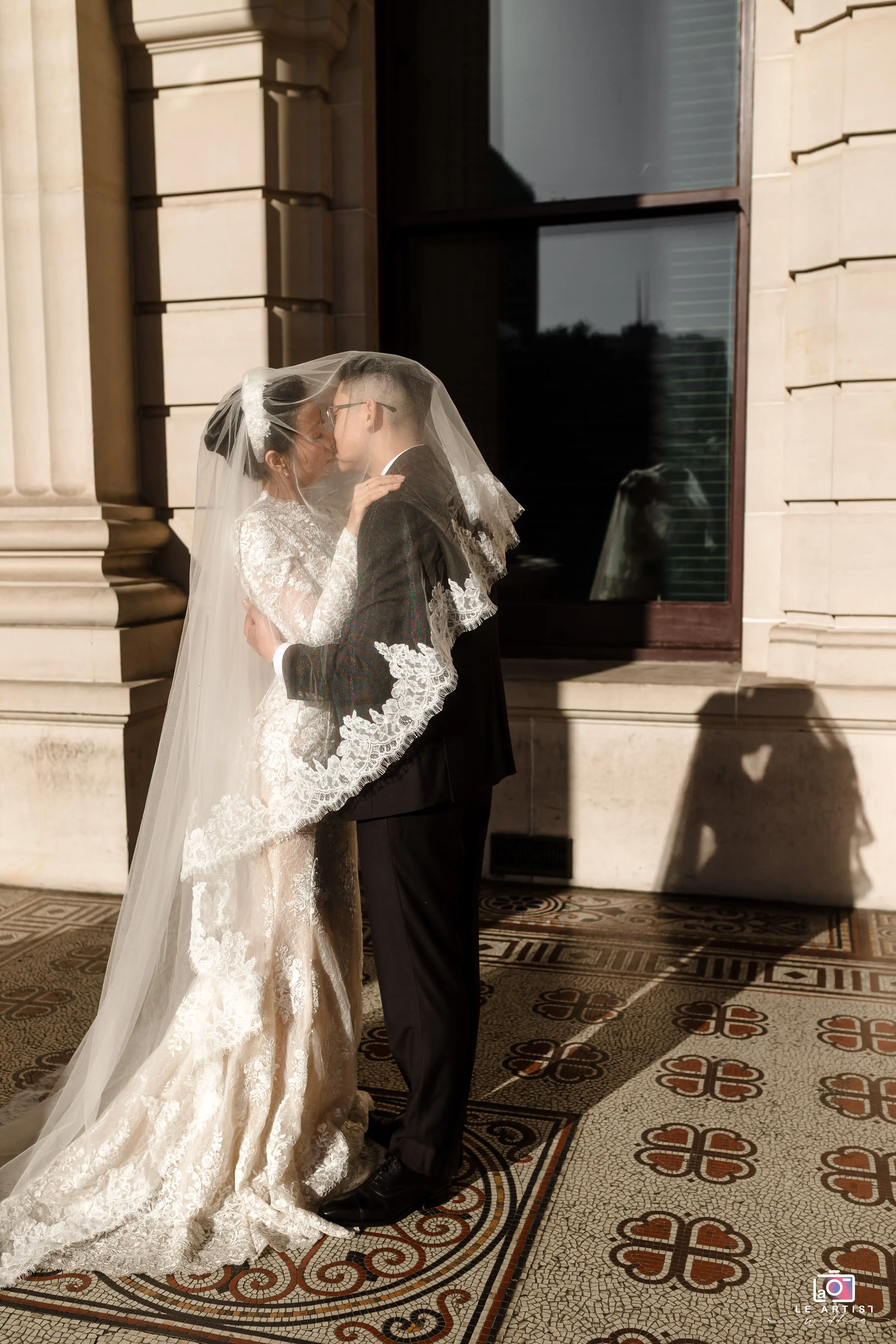 A couple shares an intimate moment under a veil on a beautifully tiled floor, casting romantic shadows against a majestic building backdrop. Le Artist Wedding