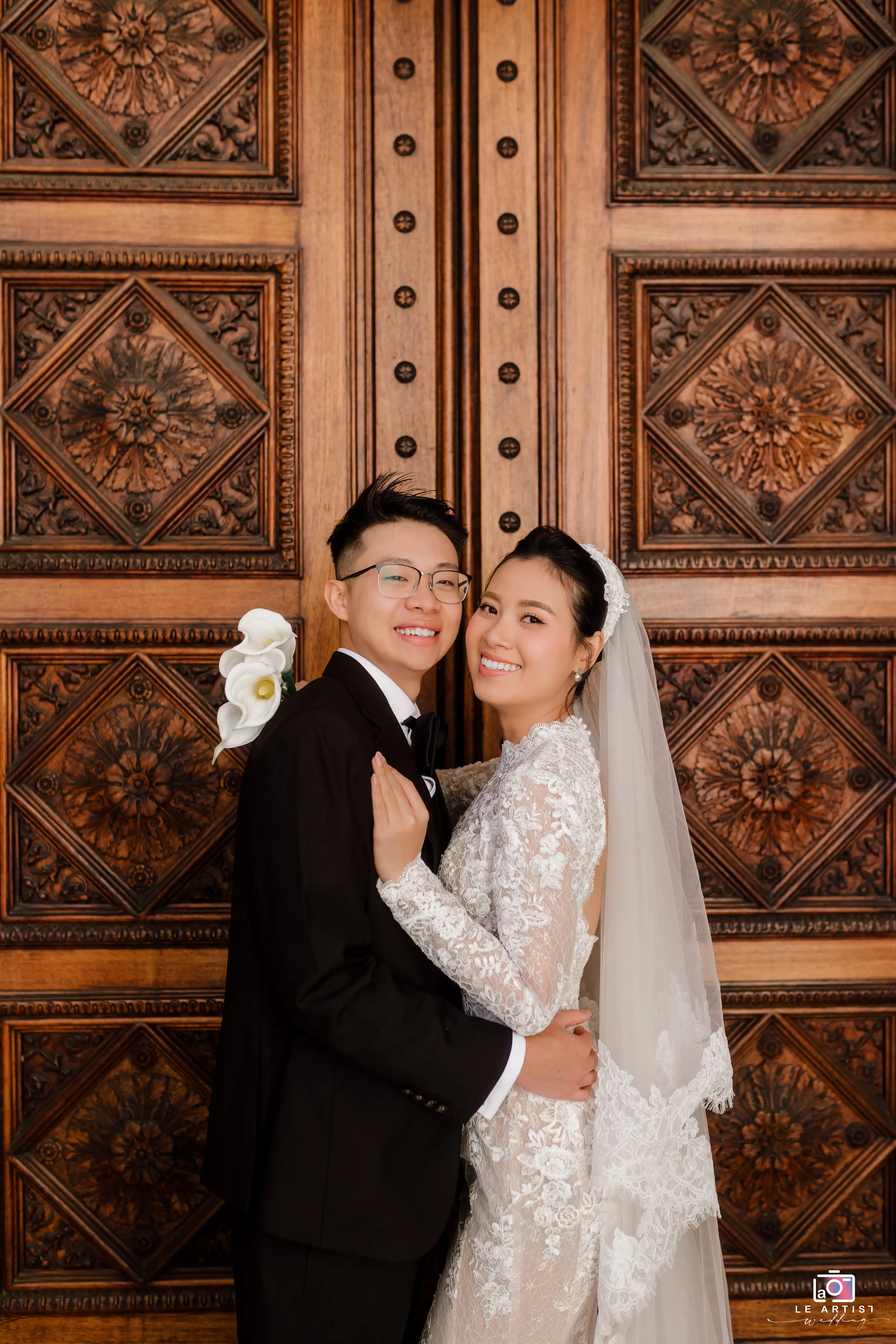 A joyful couple poses in front of an ornately carved wooden door, capturing a timeless wedding portrait. Le Artist Wedding