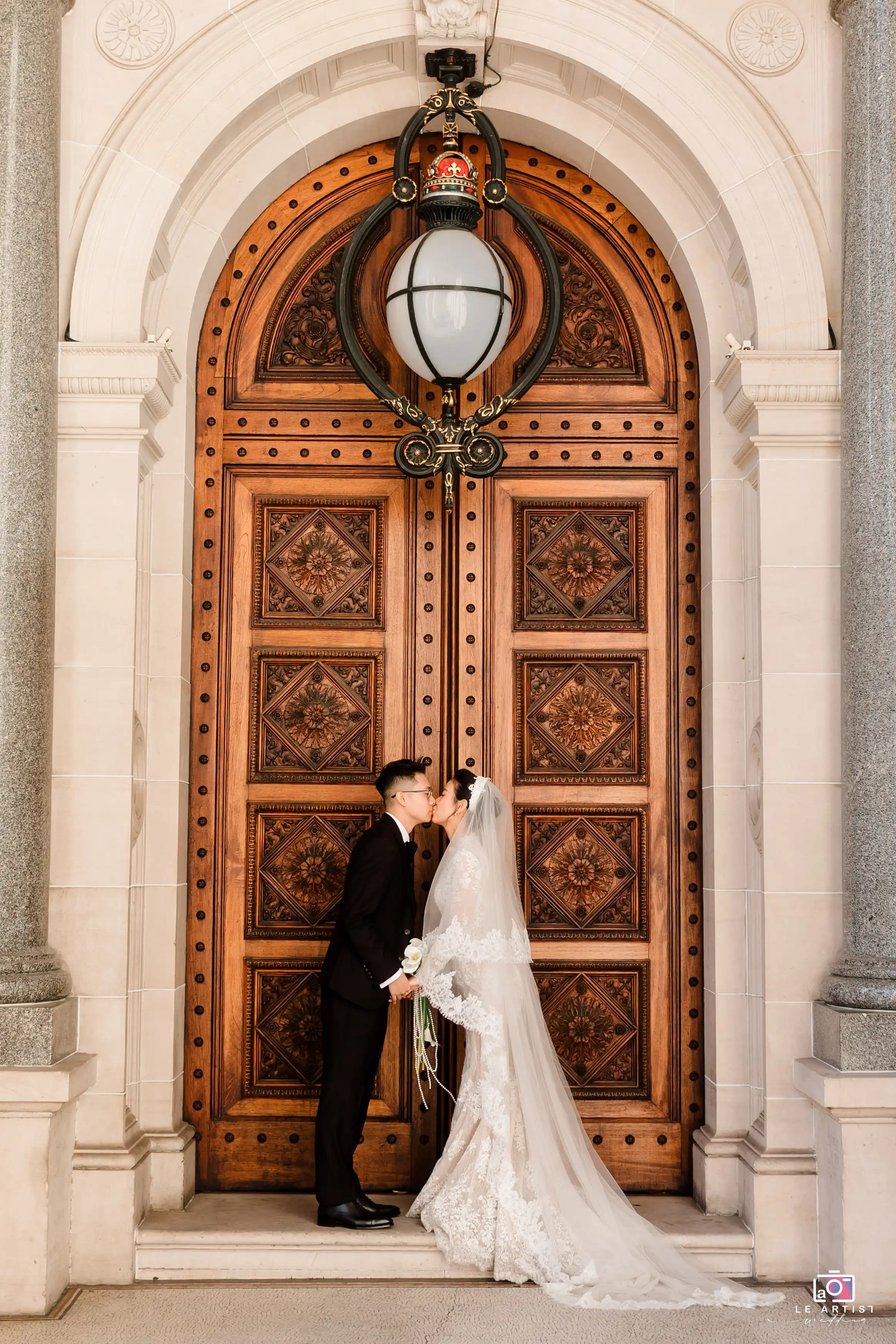 A newlywed couple shares a kiss in front of ornate wooden doors, capturing a perfect romantic moment on their wedding day. Le Artist Wedding