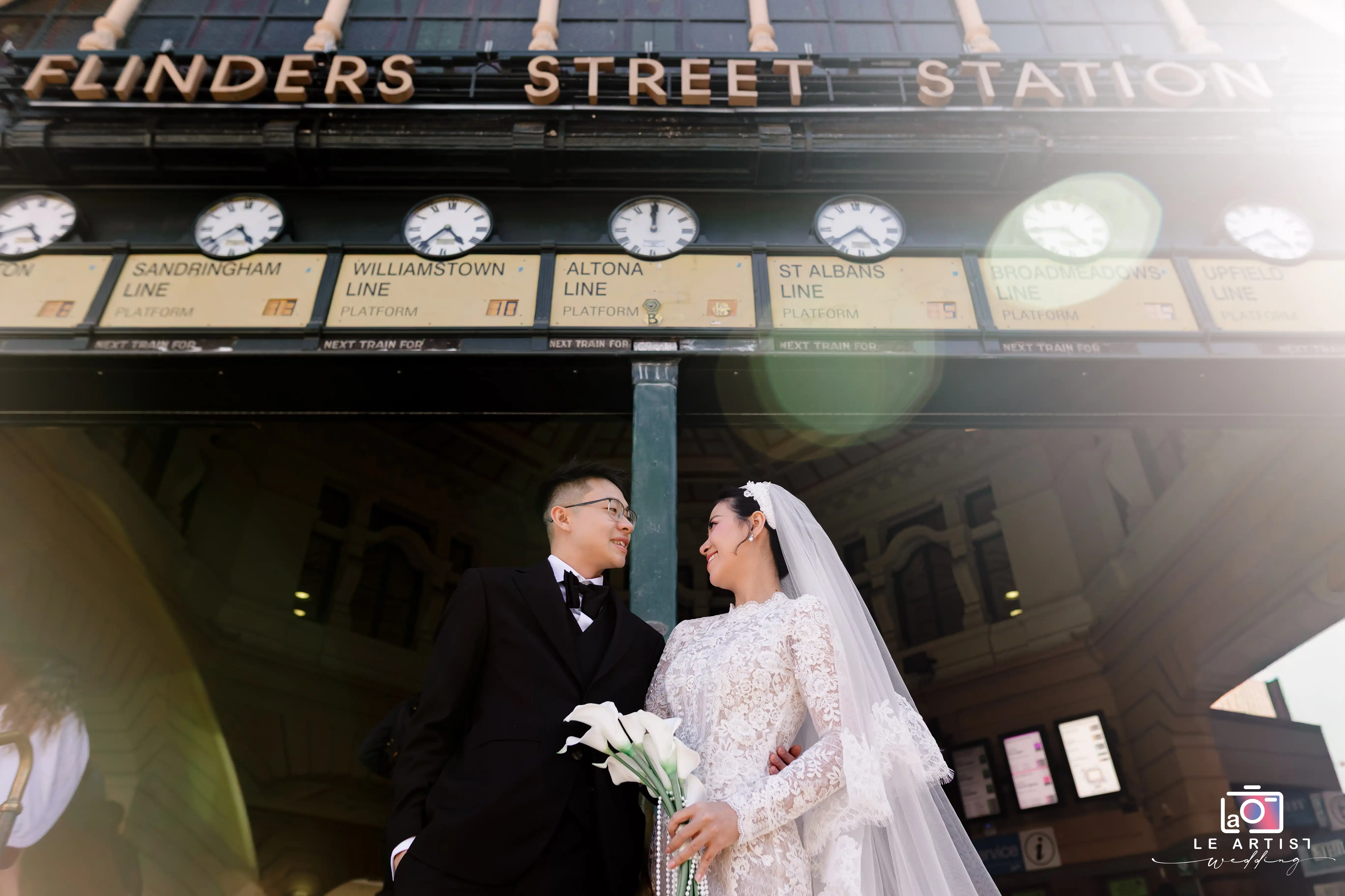 A newlywed couple shares a joyful moment outside Flinders Street Station, with the iconic clocks and architectural details providing a memorable backdrop for their special day. Le Artist Wedding