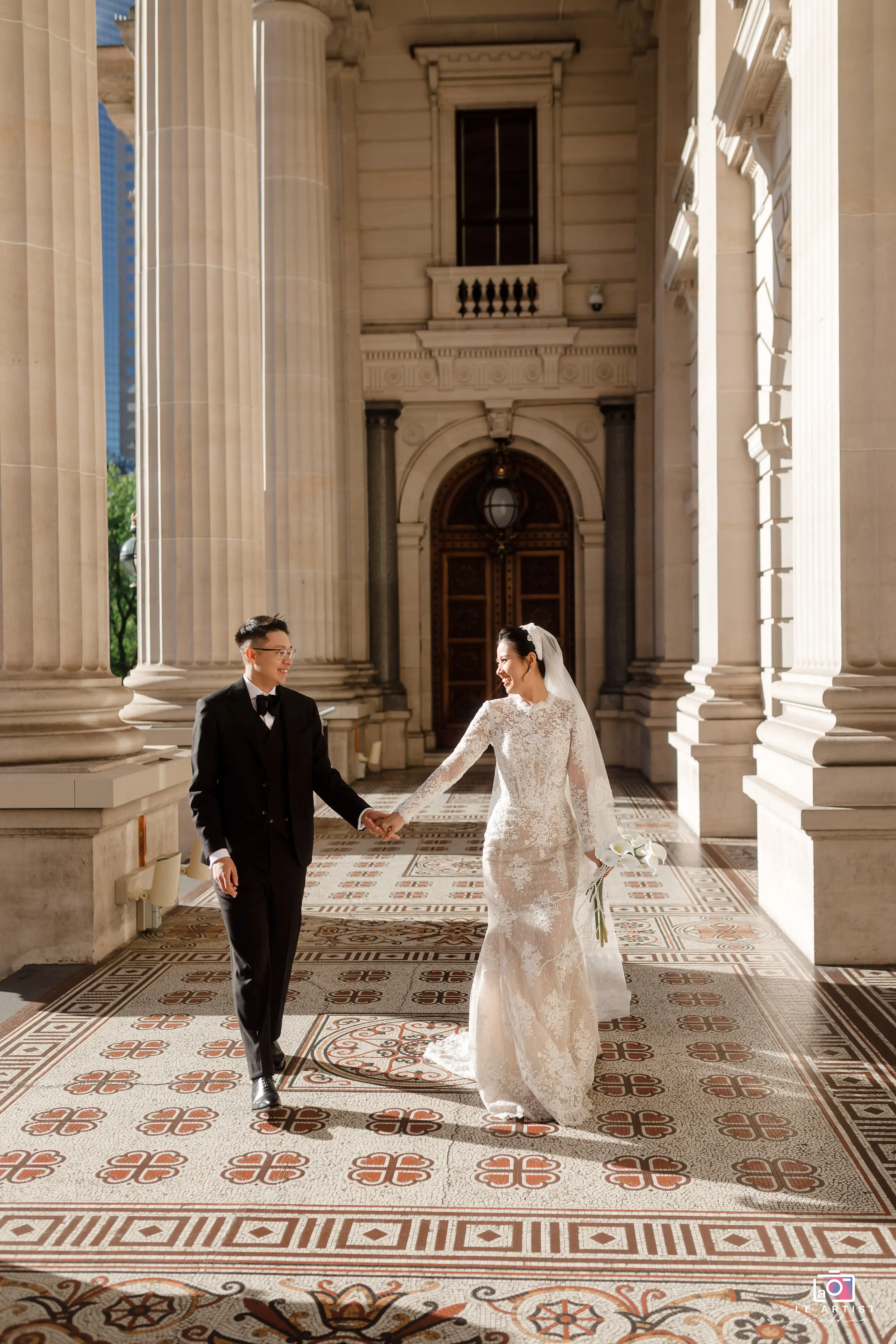 The newlyweds stroll hand in hand through a grand architectural corridor, their smiles radiant under the sunlight filtering through the columns. Le Artist Wedding