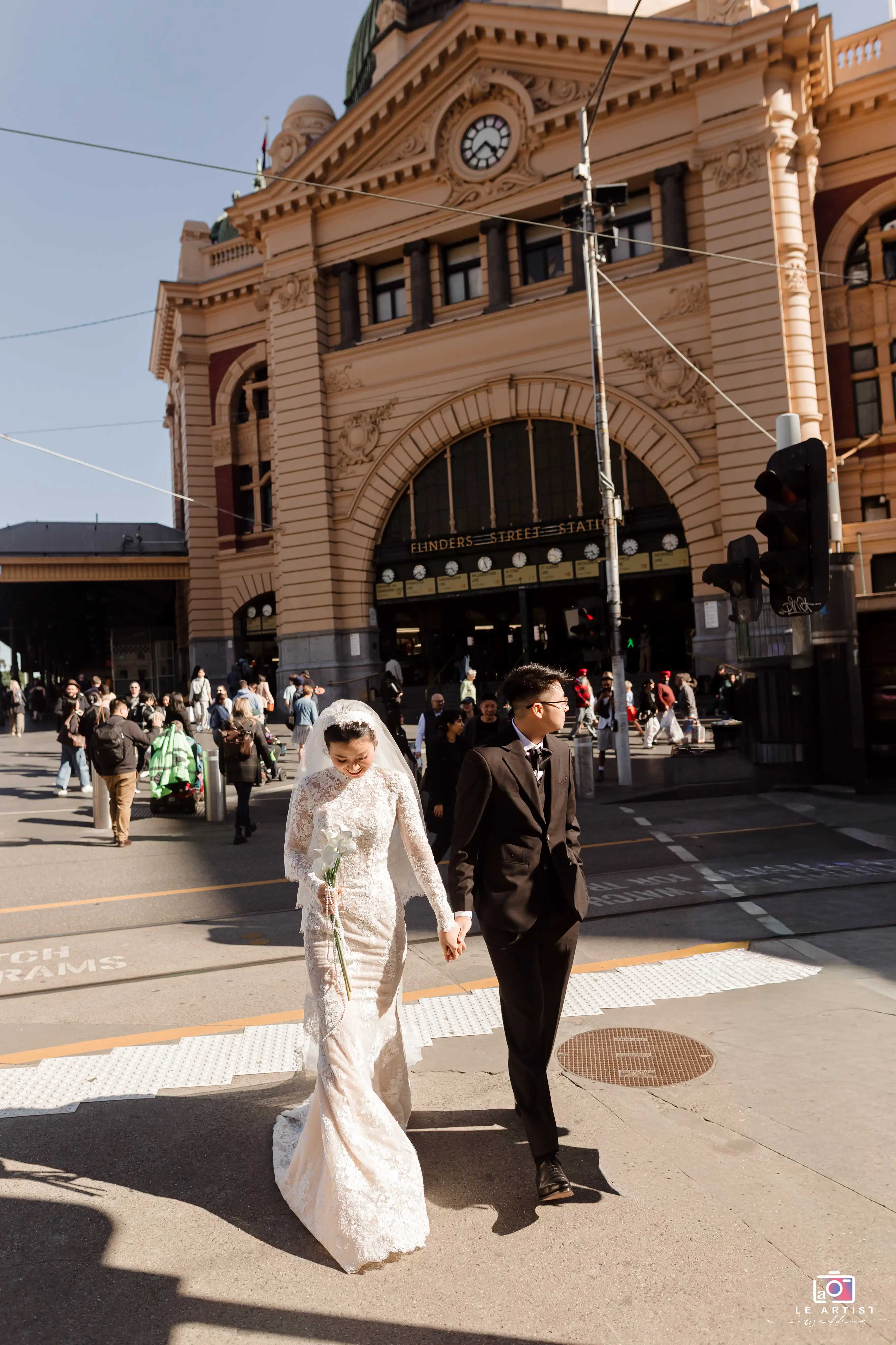 The newlywed couple stroll hand in hand outside the iconic Flinders Street Station, capturing a moment of joy and elegance during their special day. Le Artist Wedding