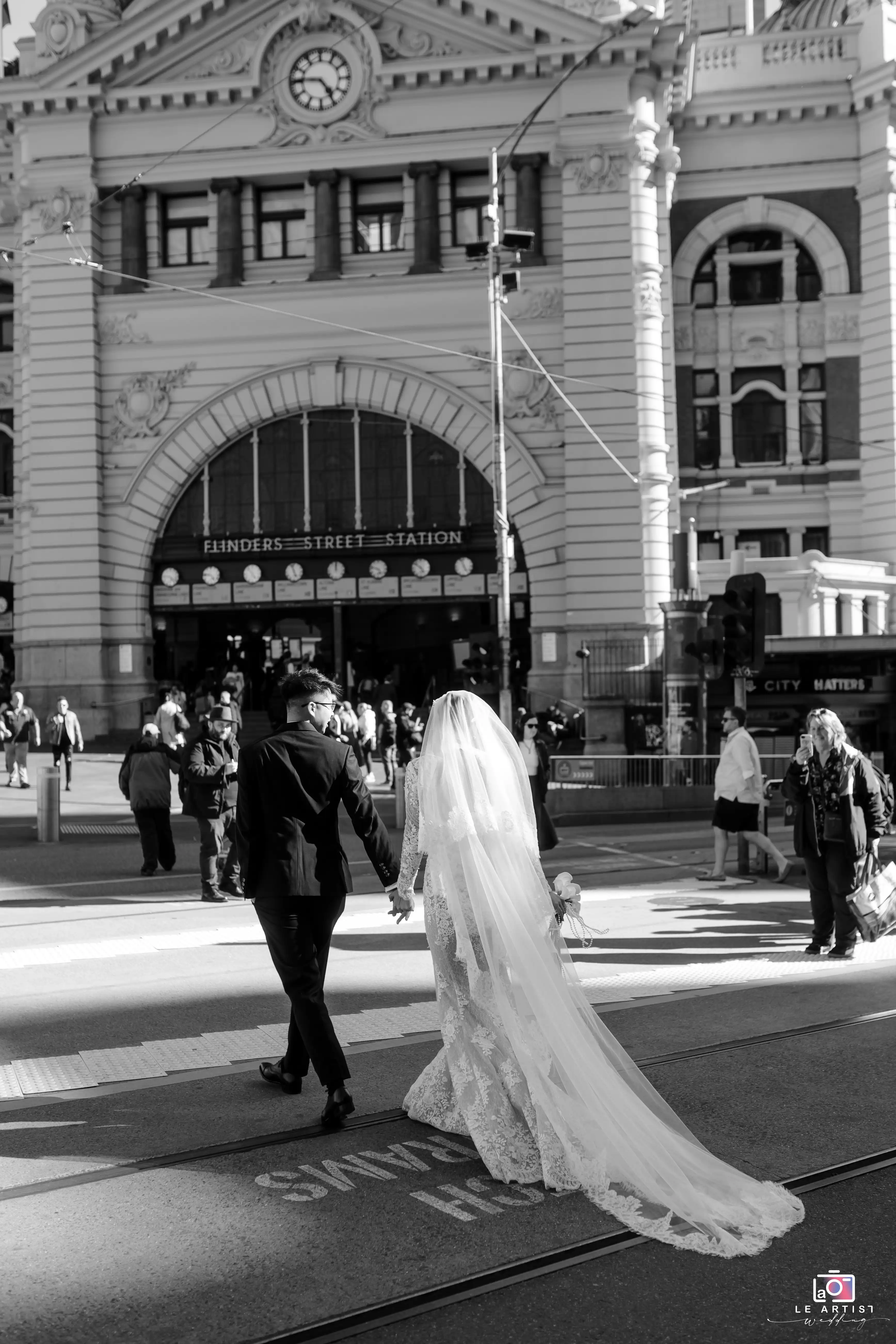 A newlywed couple walks hand in hand outside the iconic Flinders Street Station, capturing a moment of elegance and joy against the bustling city backdrop. Le Artist Wedding