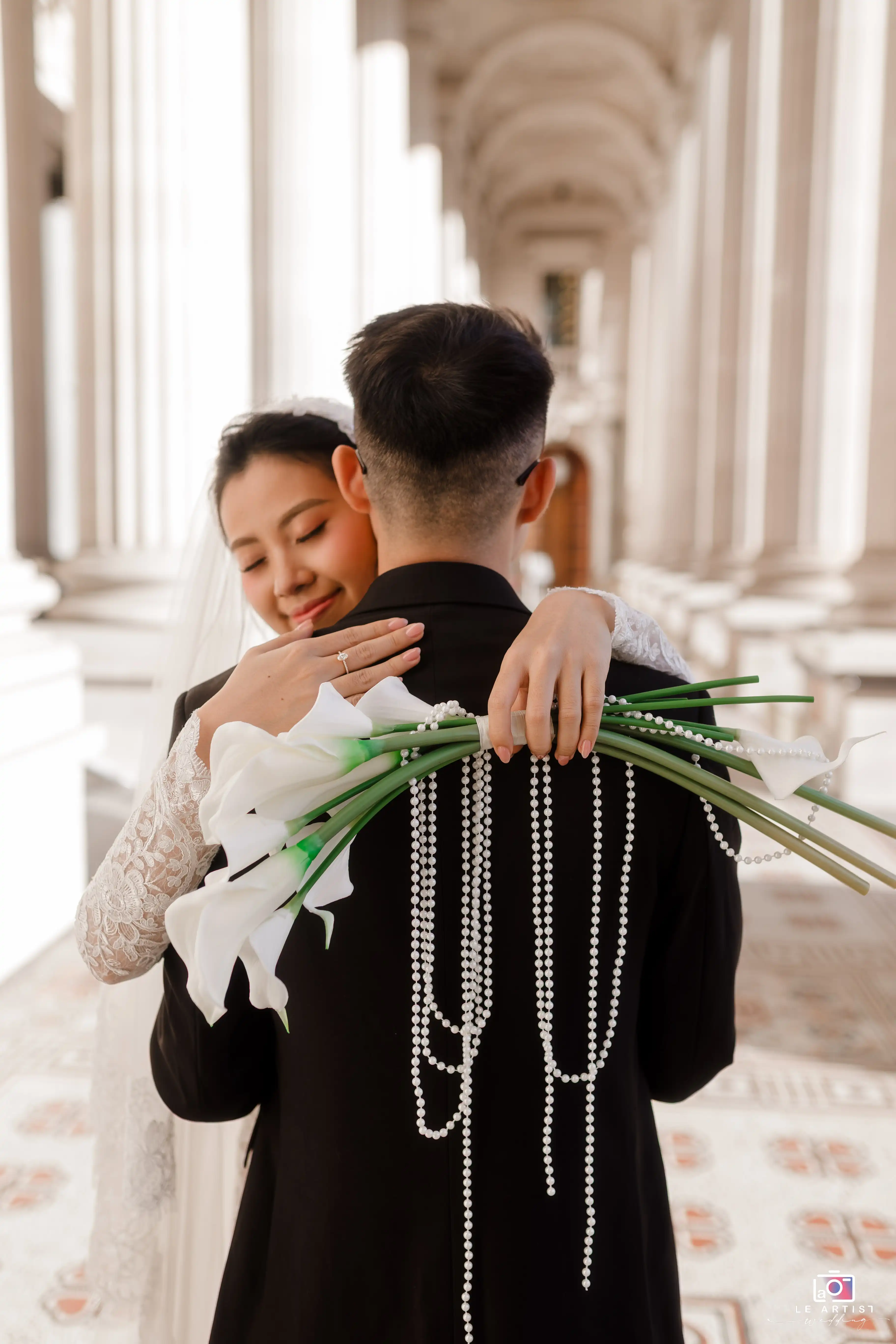 A romantic embrace between the bride and groom, with the bride holding a bouquet of calla lilies adorned with pearl strings, set against elegant architectural columns. Le Artist Wedding