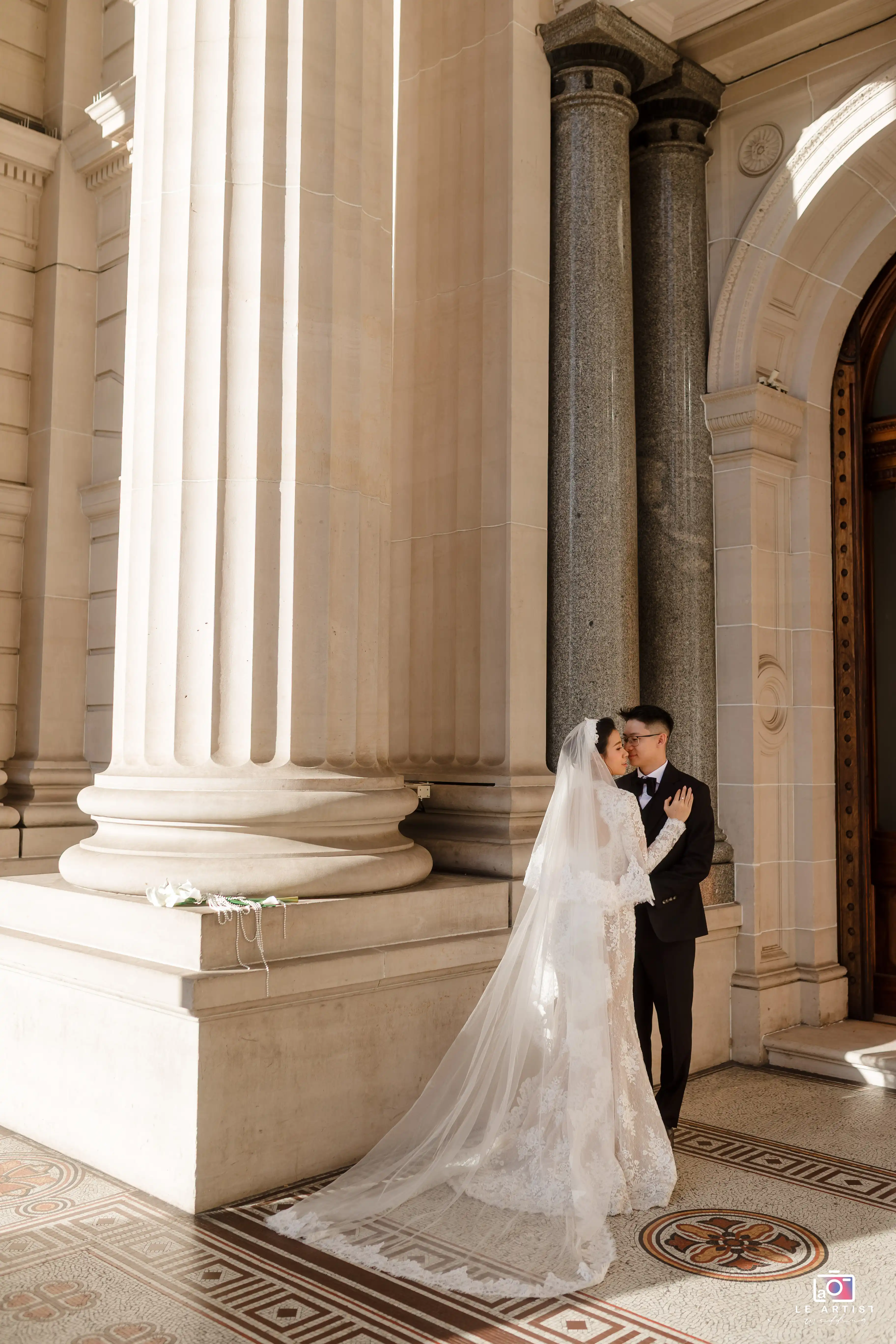 A tender moment between the newlyweds, captured in front of grand architectural columns. The bride's elegant lace gown and flowing veil add to the classic charm of the setting. Le Artist Wedding
