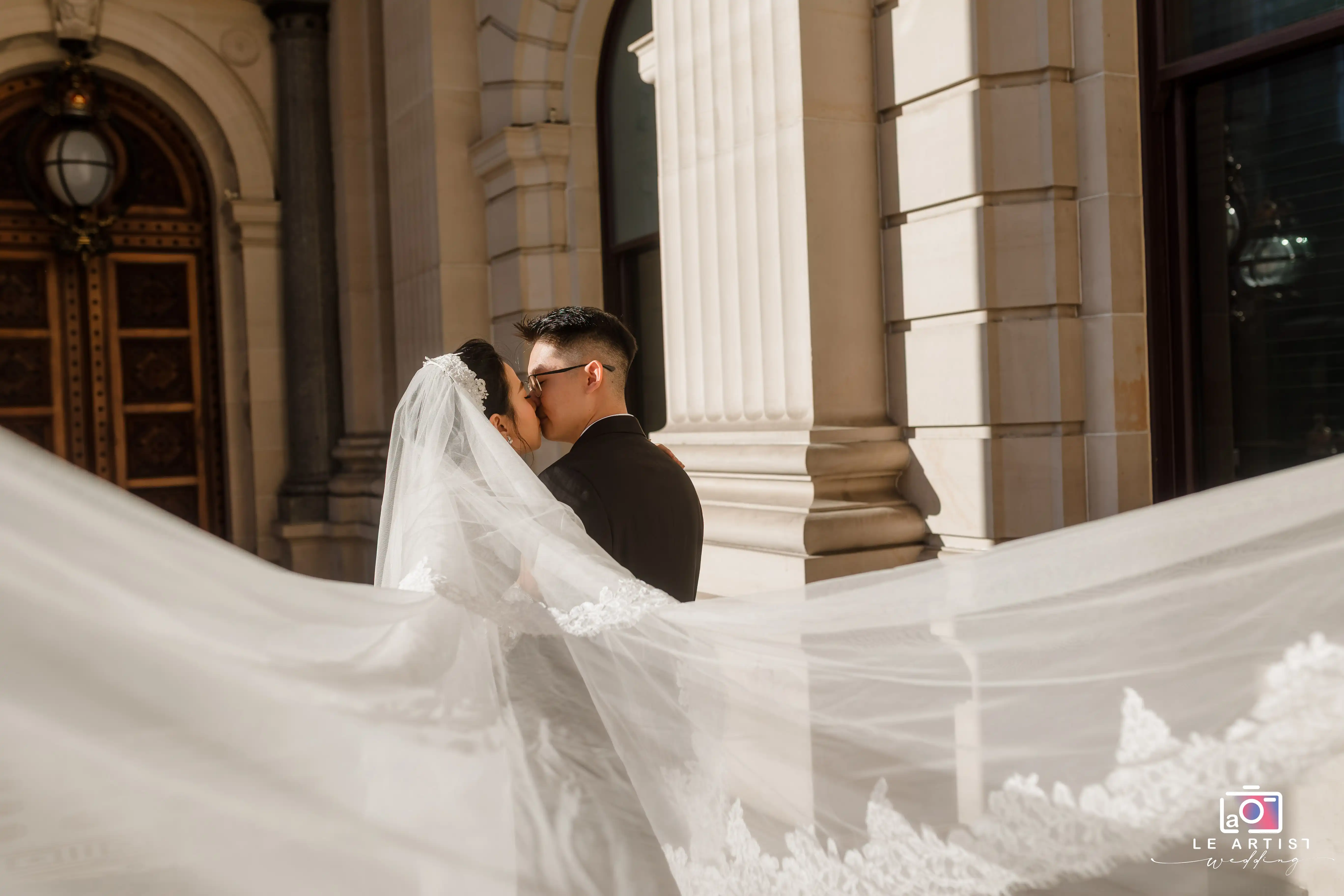 A romantic portrait of the bride and groom sharing a kiss, with the bride's veil dramatically flowing in the breeze. The elegant architecture in the background adds a touch of grandeur to the scene. Le Artist Wedding