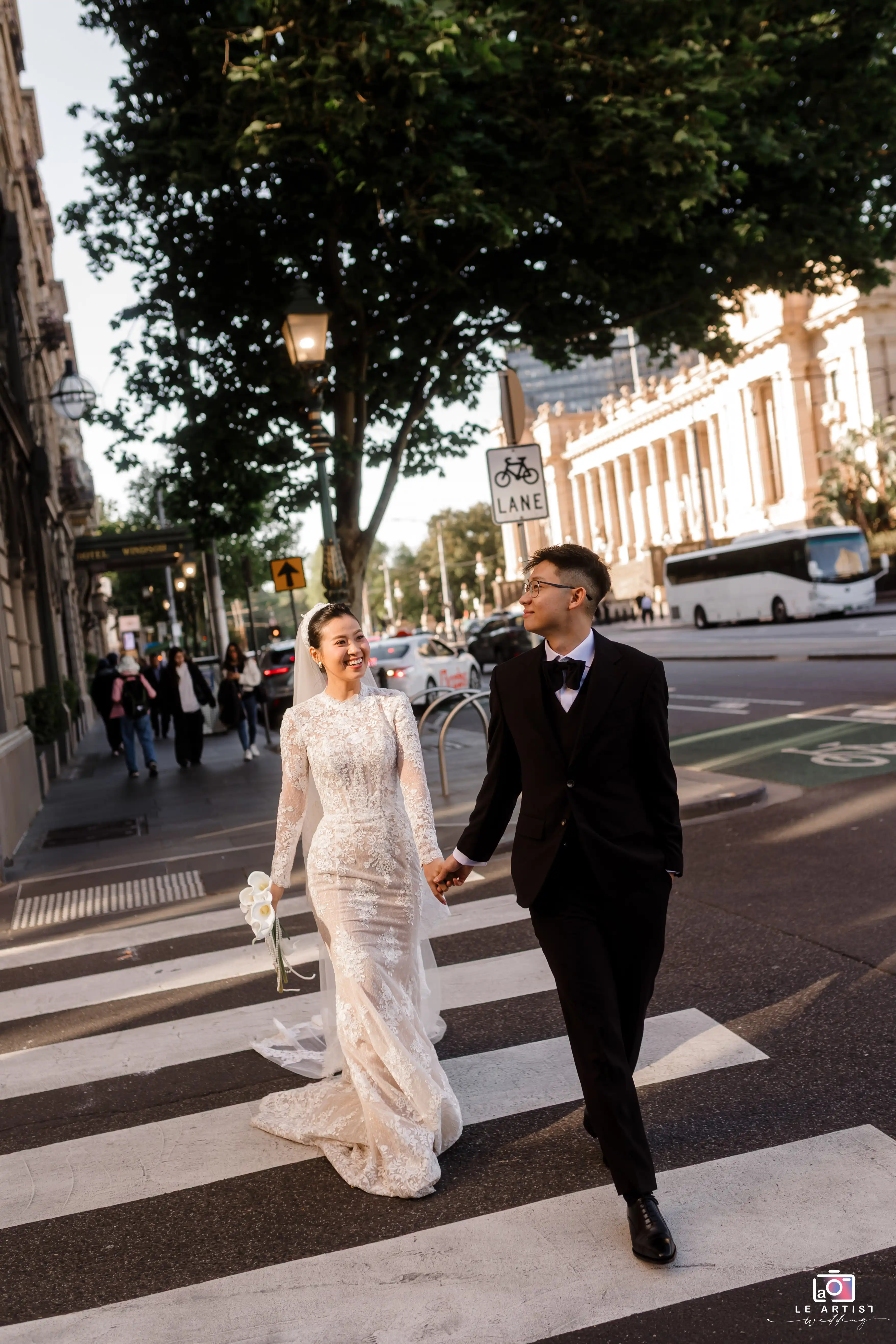 The newlyweds share a joyful moment as they walk hand in hand down a city street, capturing the essence of their special day amidst an urban backdrop. Le Artist Wedding